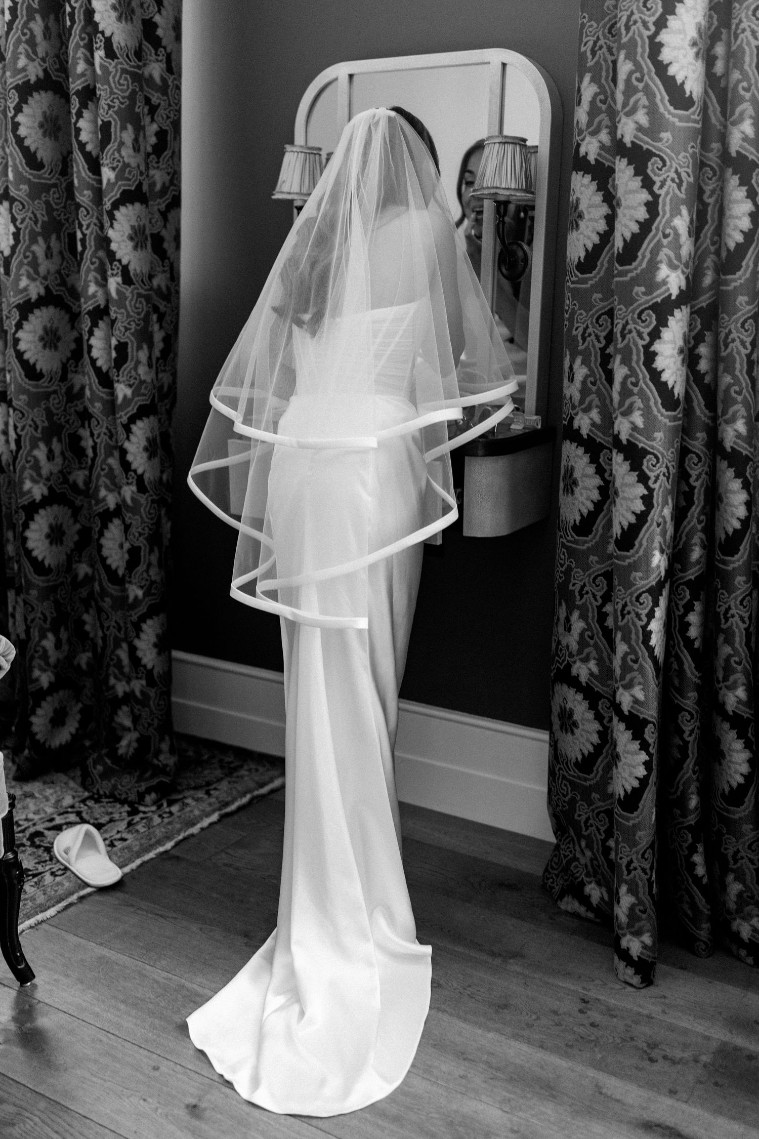 Black and white rear view of bride in elegant wedding dress and layered veil standing before a mirror.