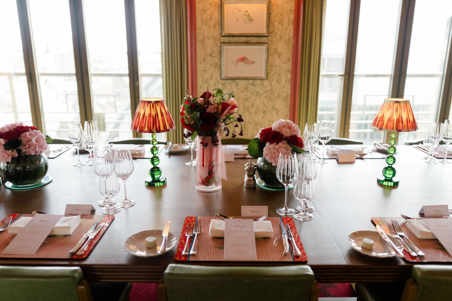 Elegant wedding reception table set with crystal glasses, pink floral centerpieces, and red lamp shades by windows.