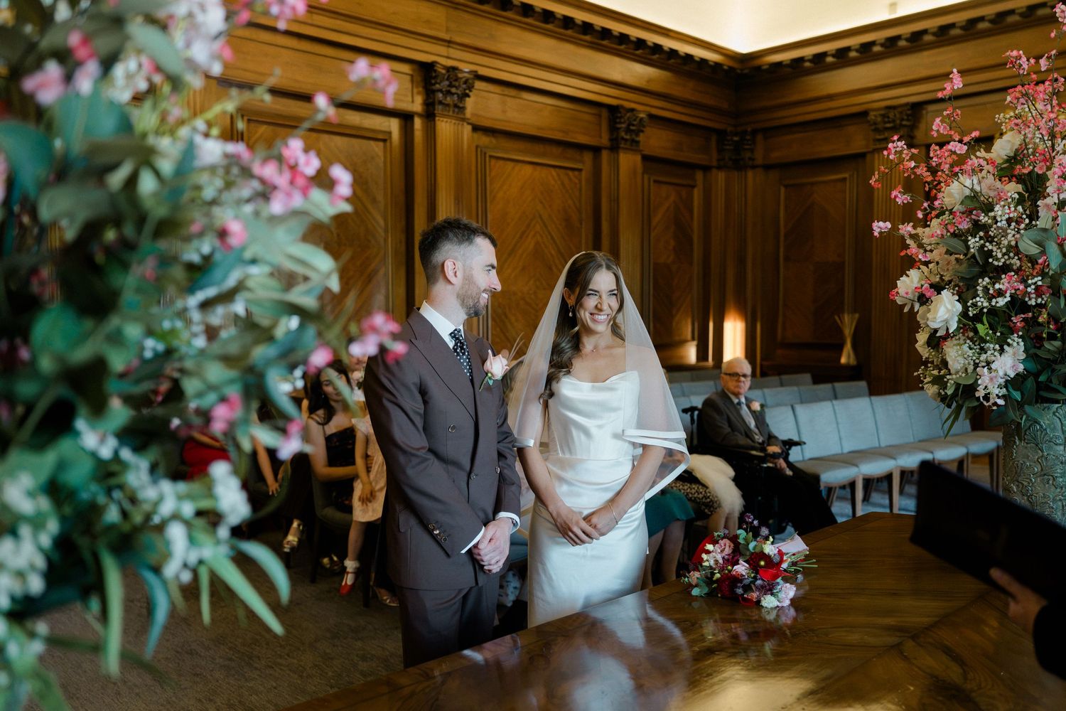 Bride and groom stand at the altar in a wood-paneled wedding ceremony room surrounded by floral arrangements.