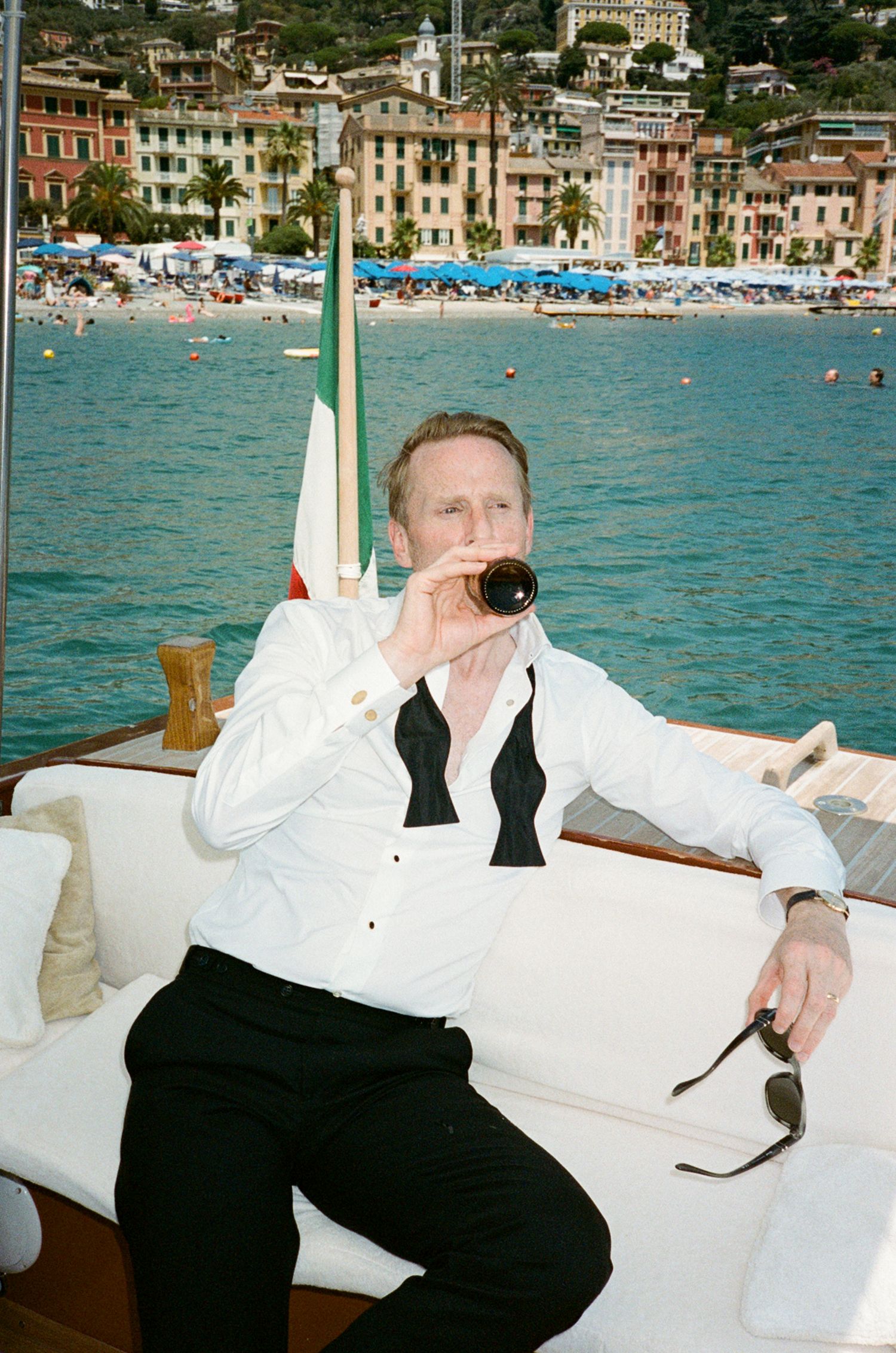Groom in white shirt and black tuxedo trousers drinks wine on a boat with colorful Italian coastal town behind him.