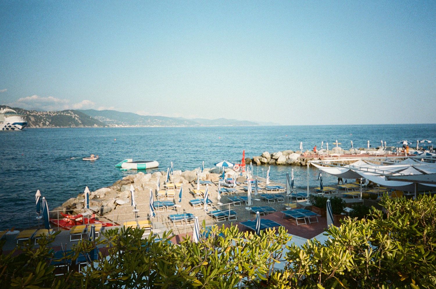 Aerial view of a sunny Mediterranean beach resort with colorful umbrellas, boats, and clear blue sea waters.