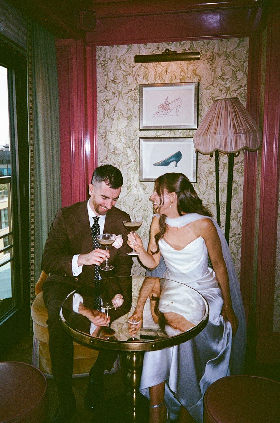 Newlyweds toast with red wine at a small bistro table in a cozy decorated room.