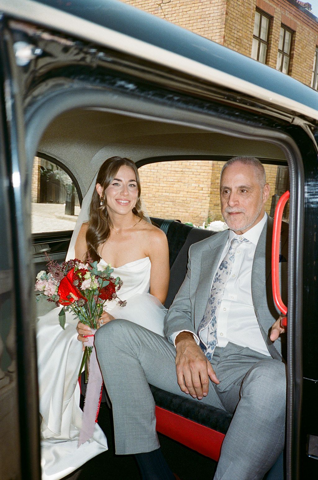 Bride in white strapless gown holding red bouquet sits with older man in gray suit inside vintage black car.