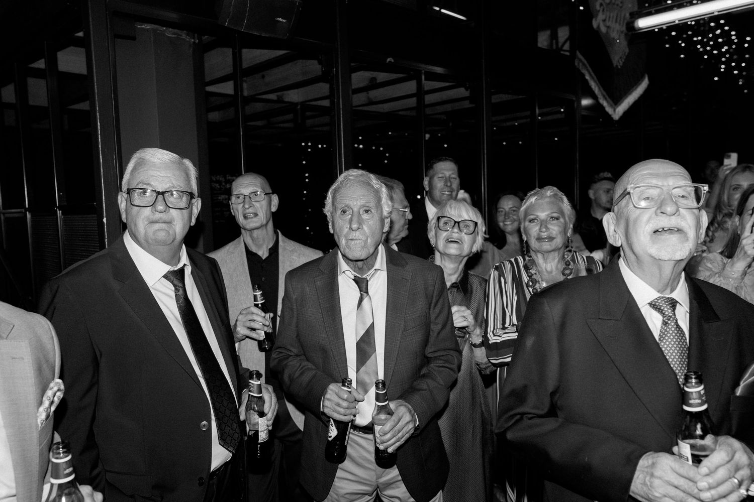 Black and white photo of elegantly dressed wedding guests holding drinks at a formal reception.