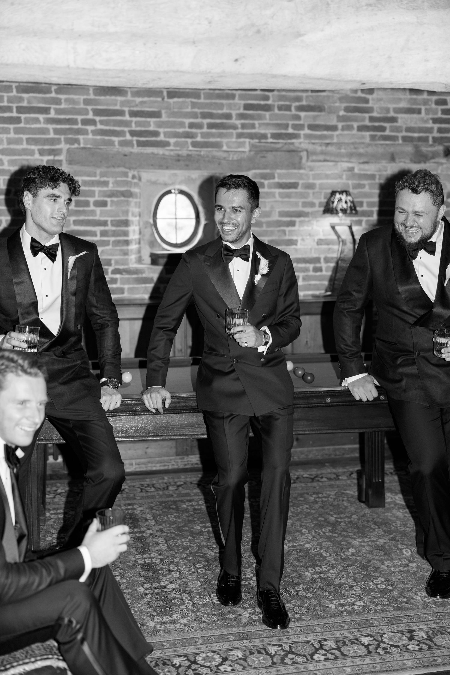 Groomsmen in black tuxedos laughing and celebrating together in a rustic brick-walled venue before the wedding.