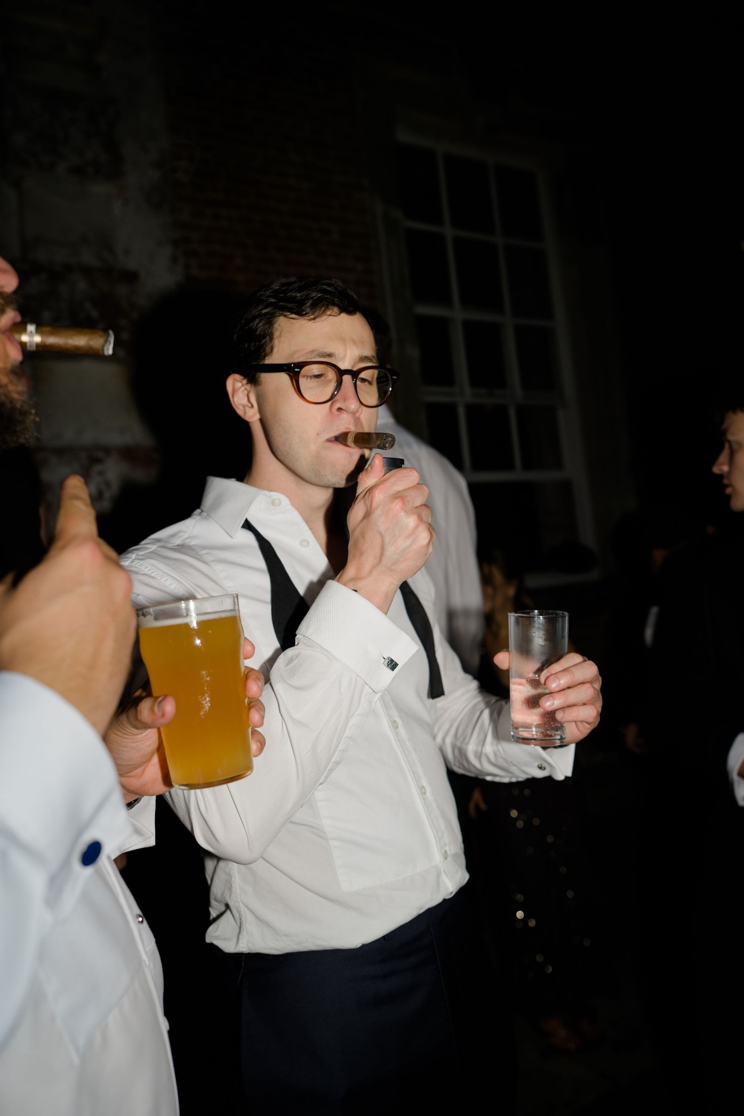 Wedding guest in glasses smokes a cigar while holding drinks at an outdoor evening reception.