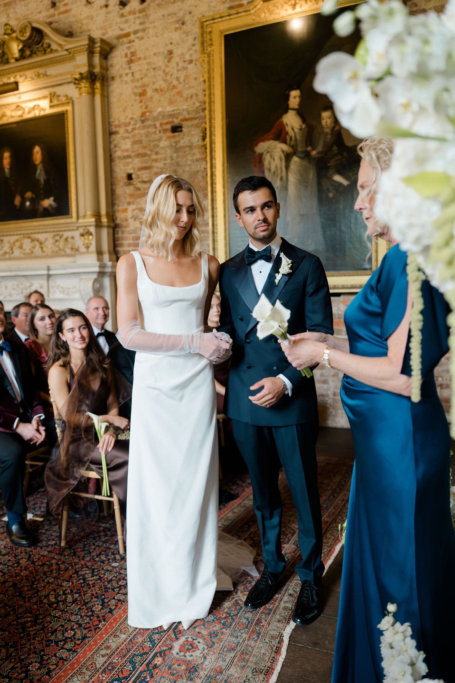 Bride in white gown and groom in navy tuxedo exchange vows with officiant in elegant historic venue.