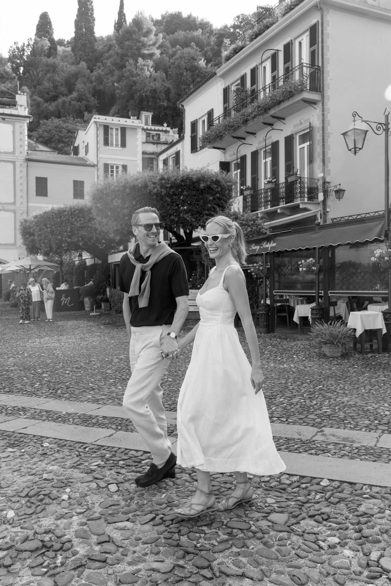 Elegant couple strolling hand in hand through charming Italian cobblestone piazza, black and white photography.