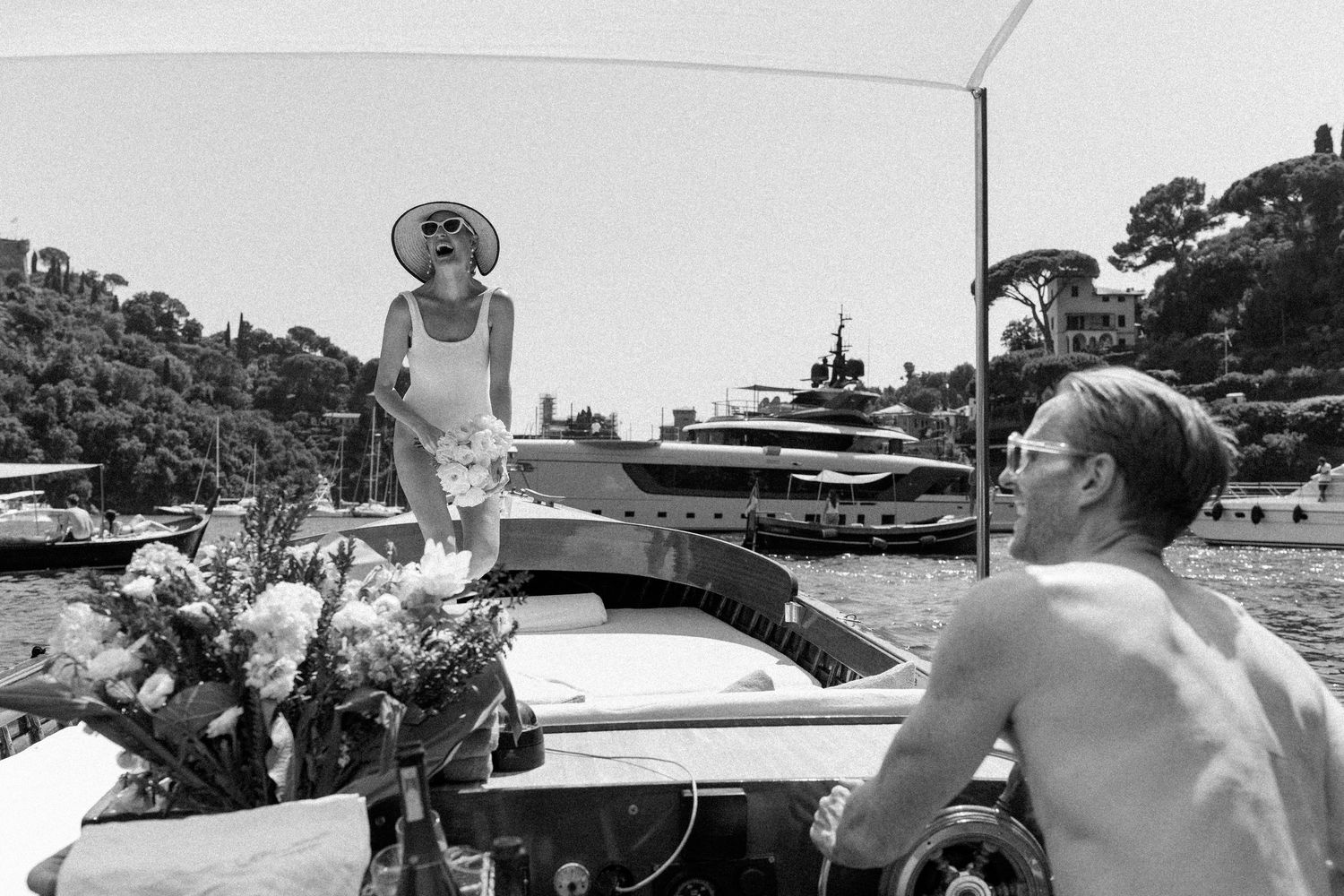 Woman in white swimsuit and sun hat standing on a luxury yacht in a scenic Mediterranean harbor.