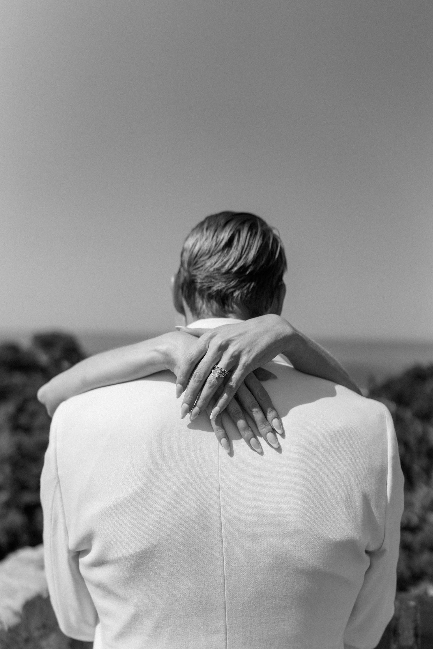 Bride places ringed hand on grooms white suit jacket back with ocean view behind them.