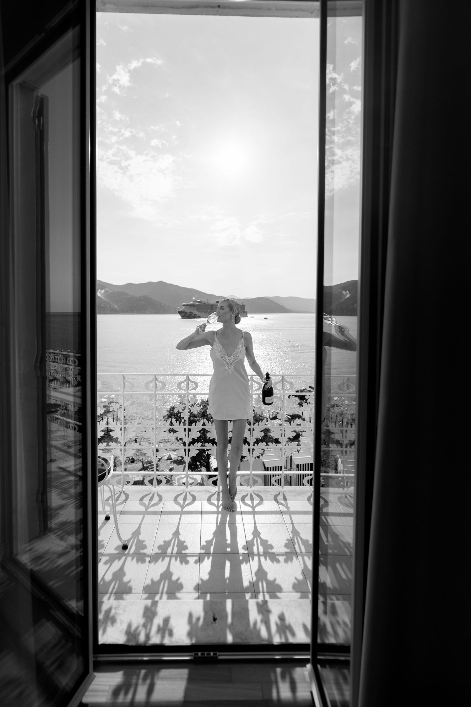 Black and white photo of a bride in a short dress standing on a sunlit balcony overlooking the sea.