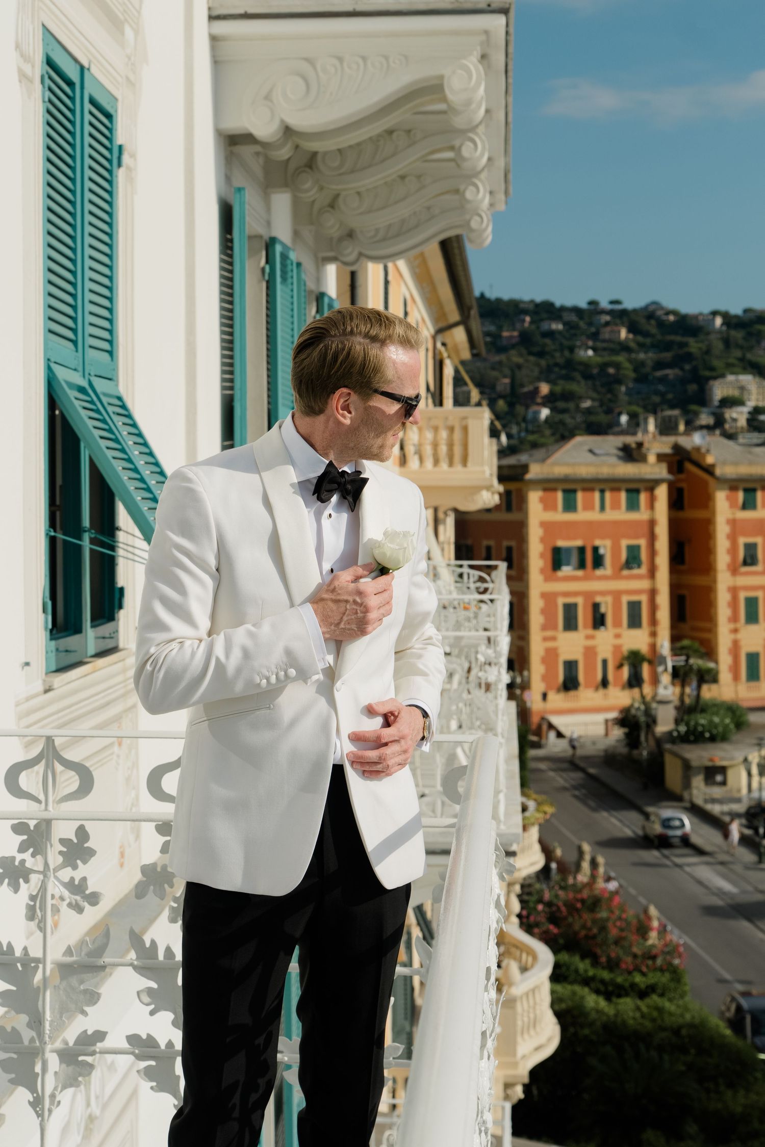 Groom in white tuxedo jacket holding boutonniere on ornate balcony overlooking Italian town.