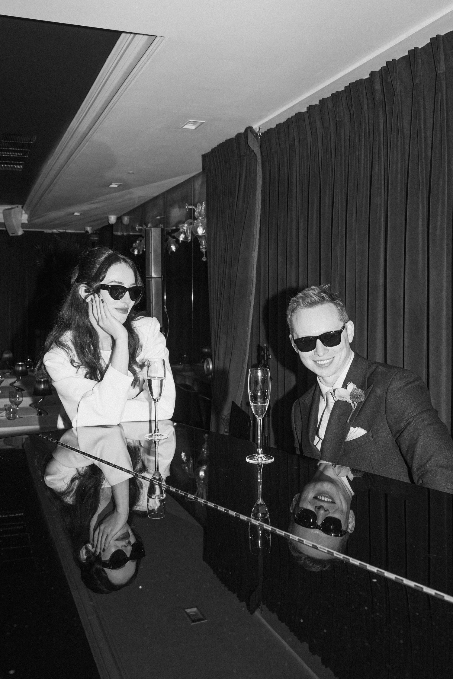 Black and white photo of newlywed couple wearing sunglasses celebrating with champagne at bar.