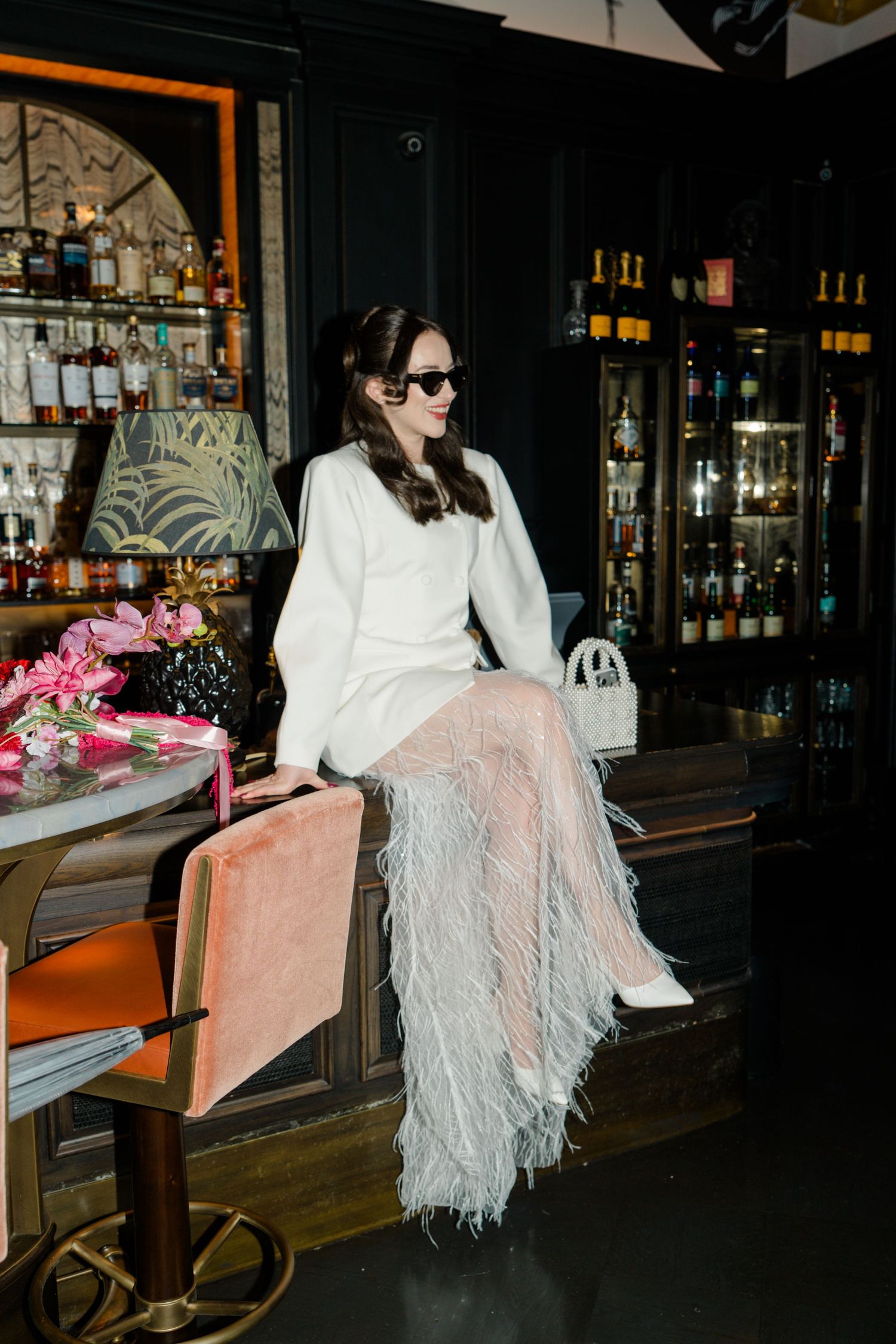 Stylish bride in white blazer and feathered skirt sitting at dark bar wearing sunglasses with pink bouquet.