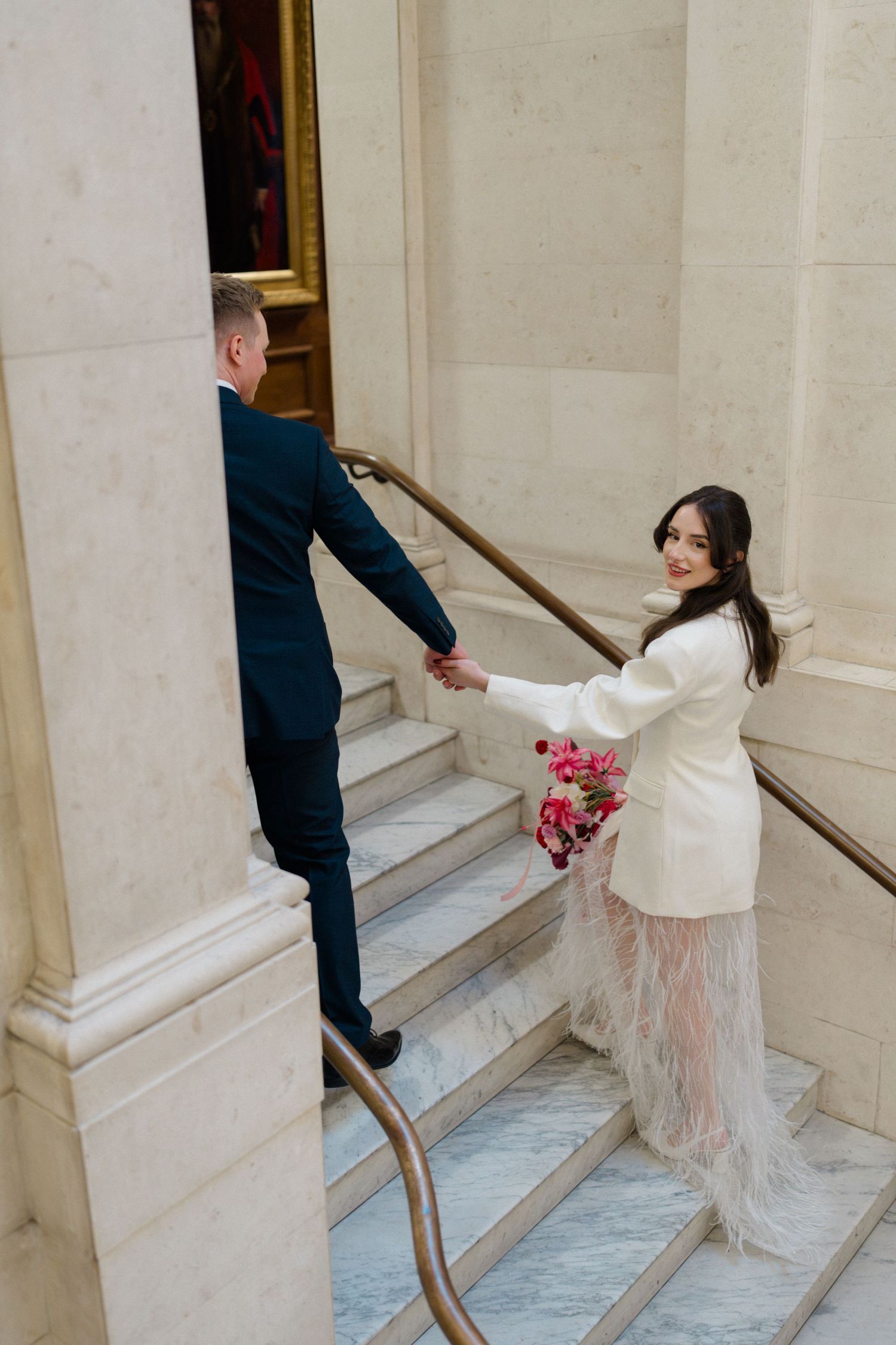 Bride in a white blazer and feather skirt holding hands with groom on marble wedding venue stairs.