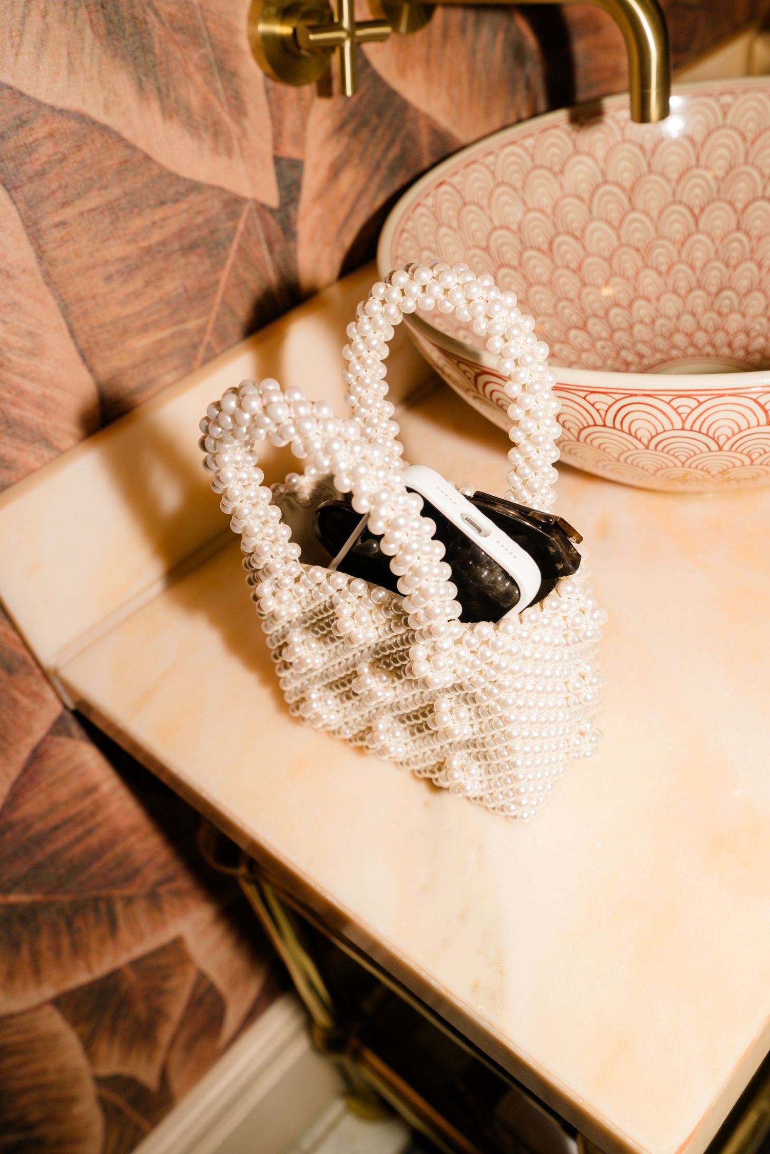 Pearl beaded bridal handbag on marble vanity beside pink scalloped sink with gold fixtures.