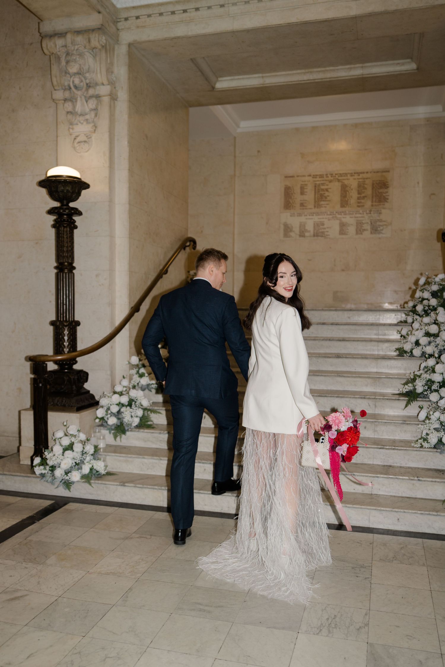 Bride in white coat dress with lace train holds bouquet ascending marble stairs with groom in navy suit.