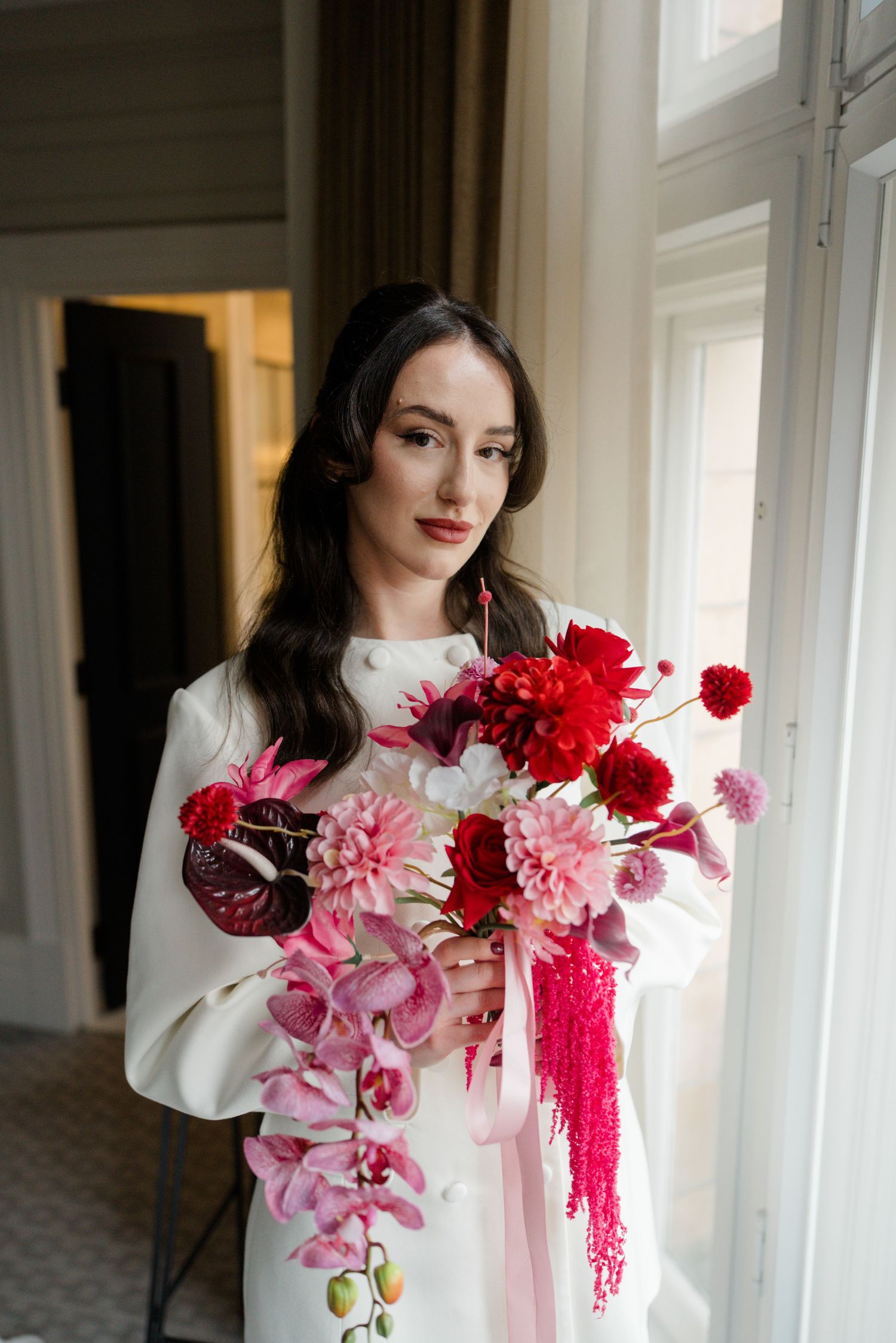 Bride in white coat holding vibrant red and pink floral bouquet beside tall windows.