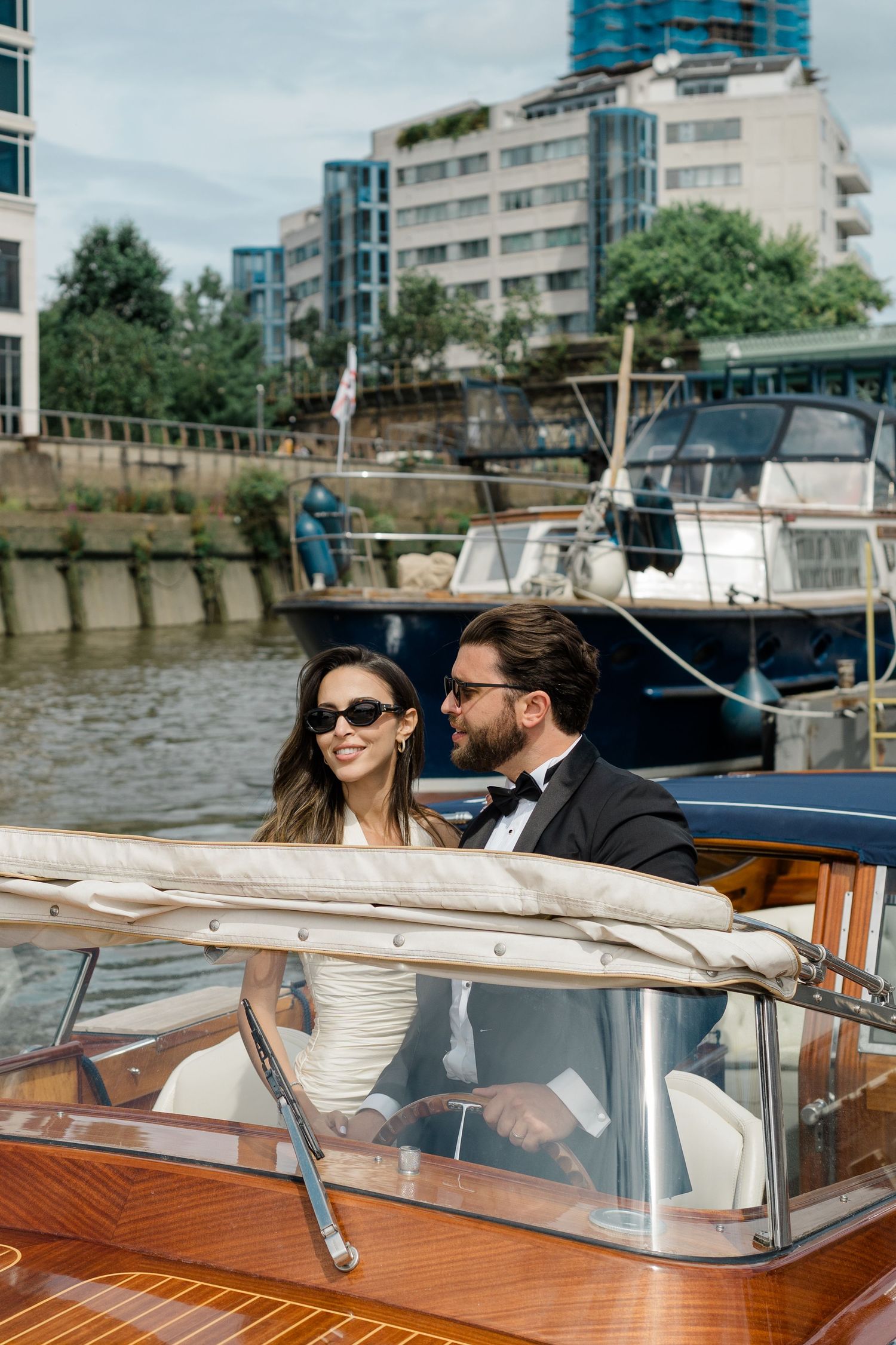 Elegant couple in sunglasses riding a classic wooden boat through a marina with yachts and city buildings in background.