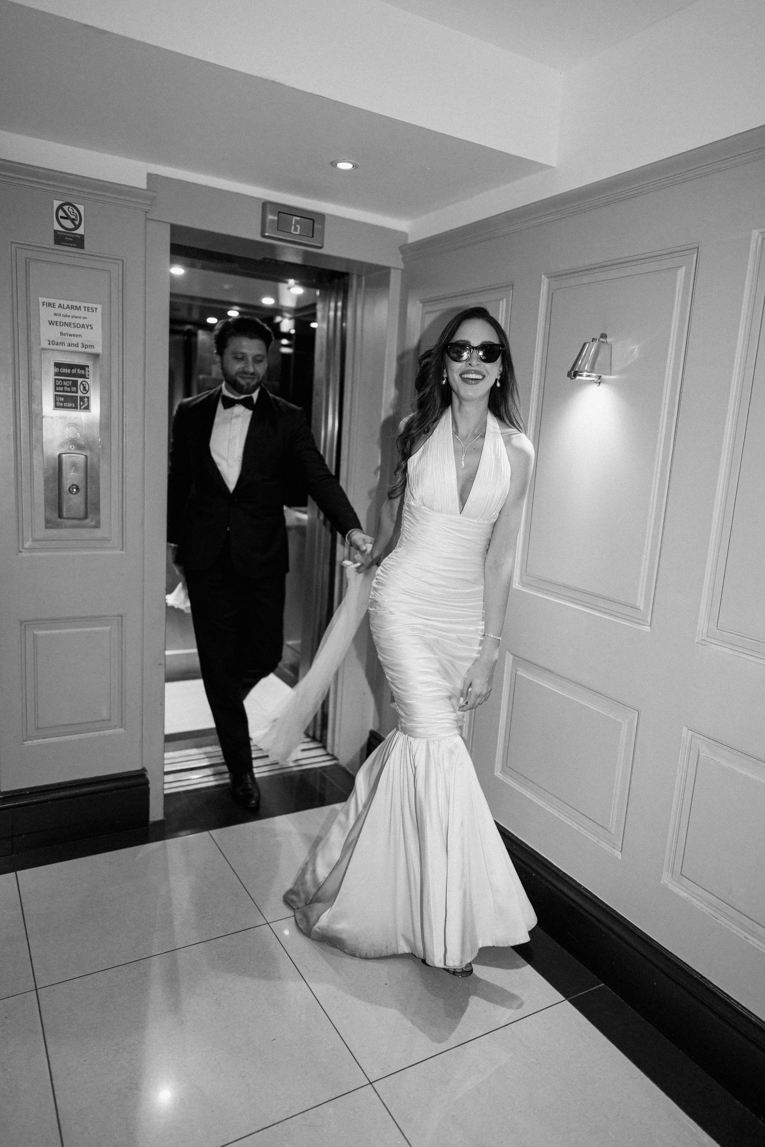 Bride in white mermaid gown and sunglasses exits elevator with groom in tuxedo, black and white wedding photo.