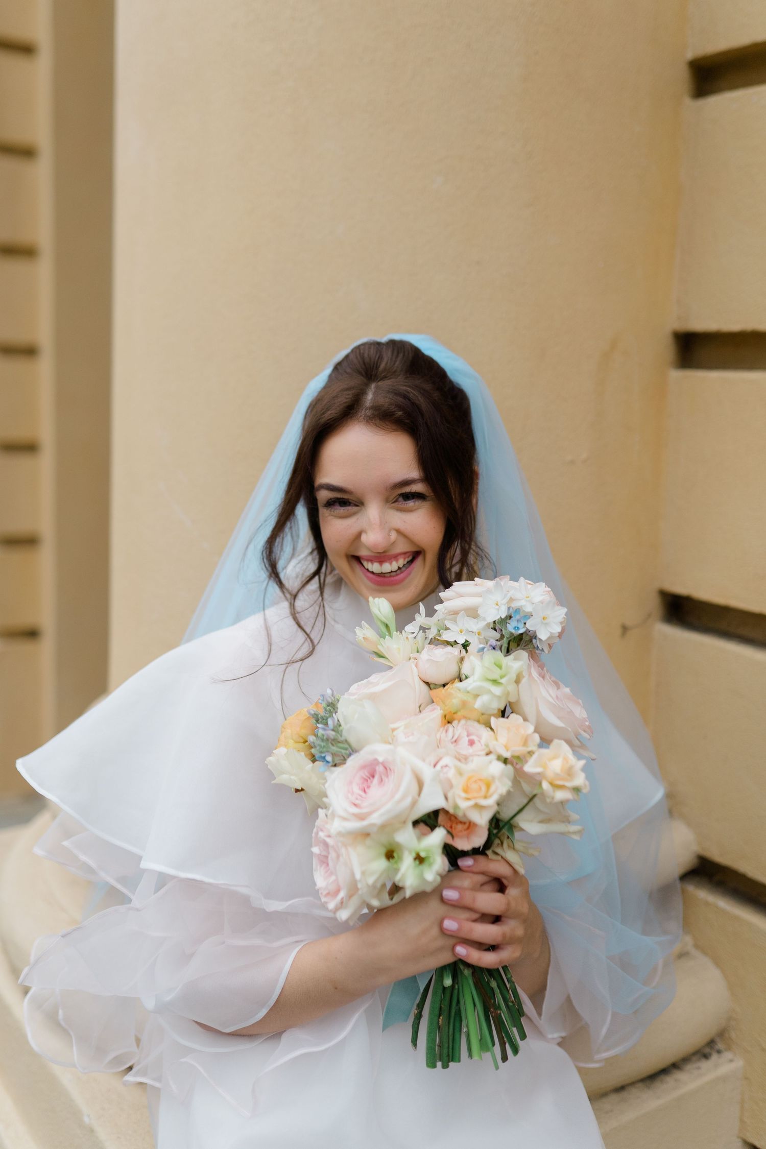 Smiling bride in white dress and blue veil holding a pastel bouquet of roses outdoors.