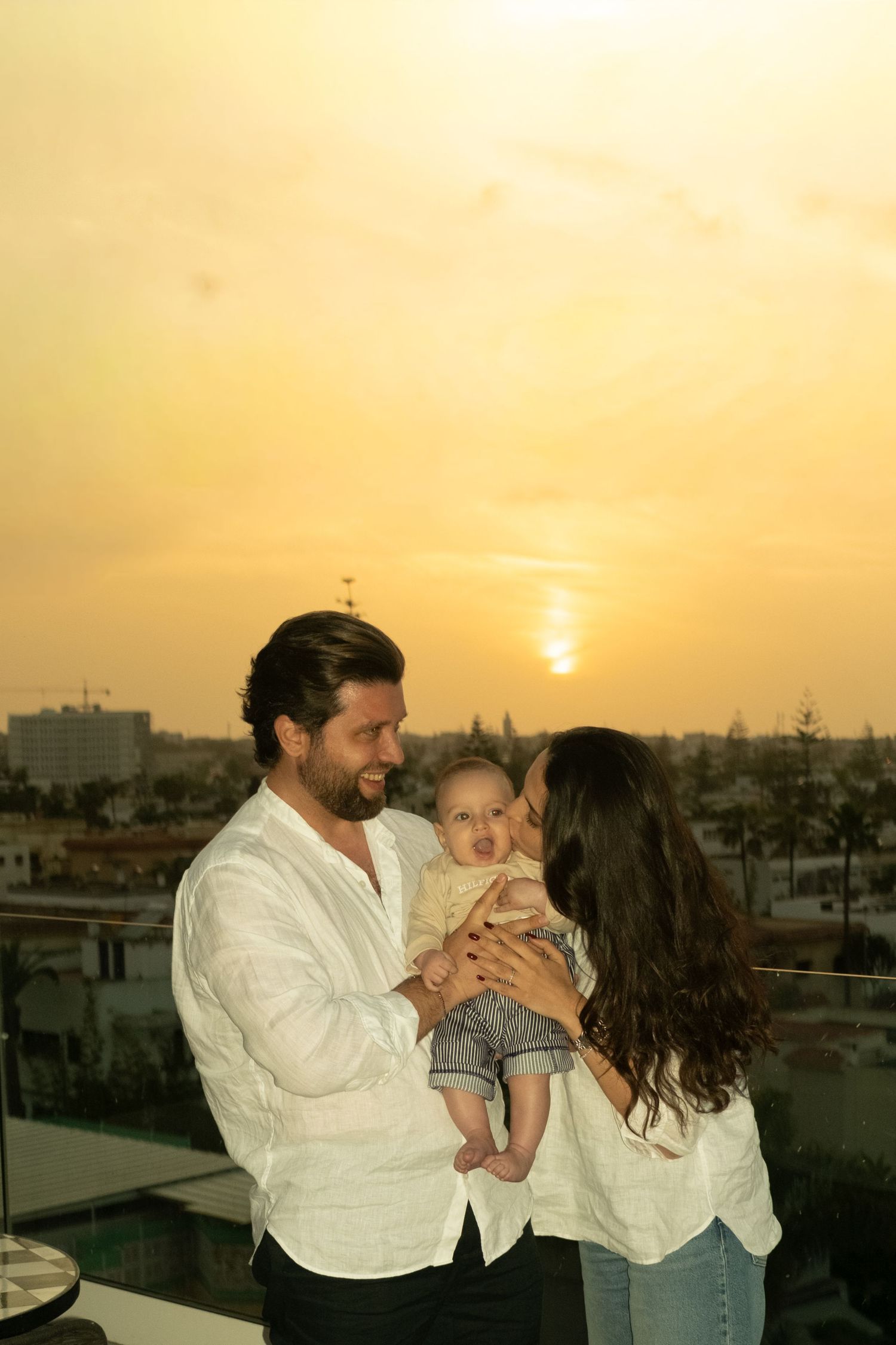 A couple shares a tender moment with their baby on a rooftop terrace during a golden sunset over the city.