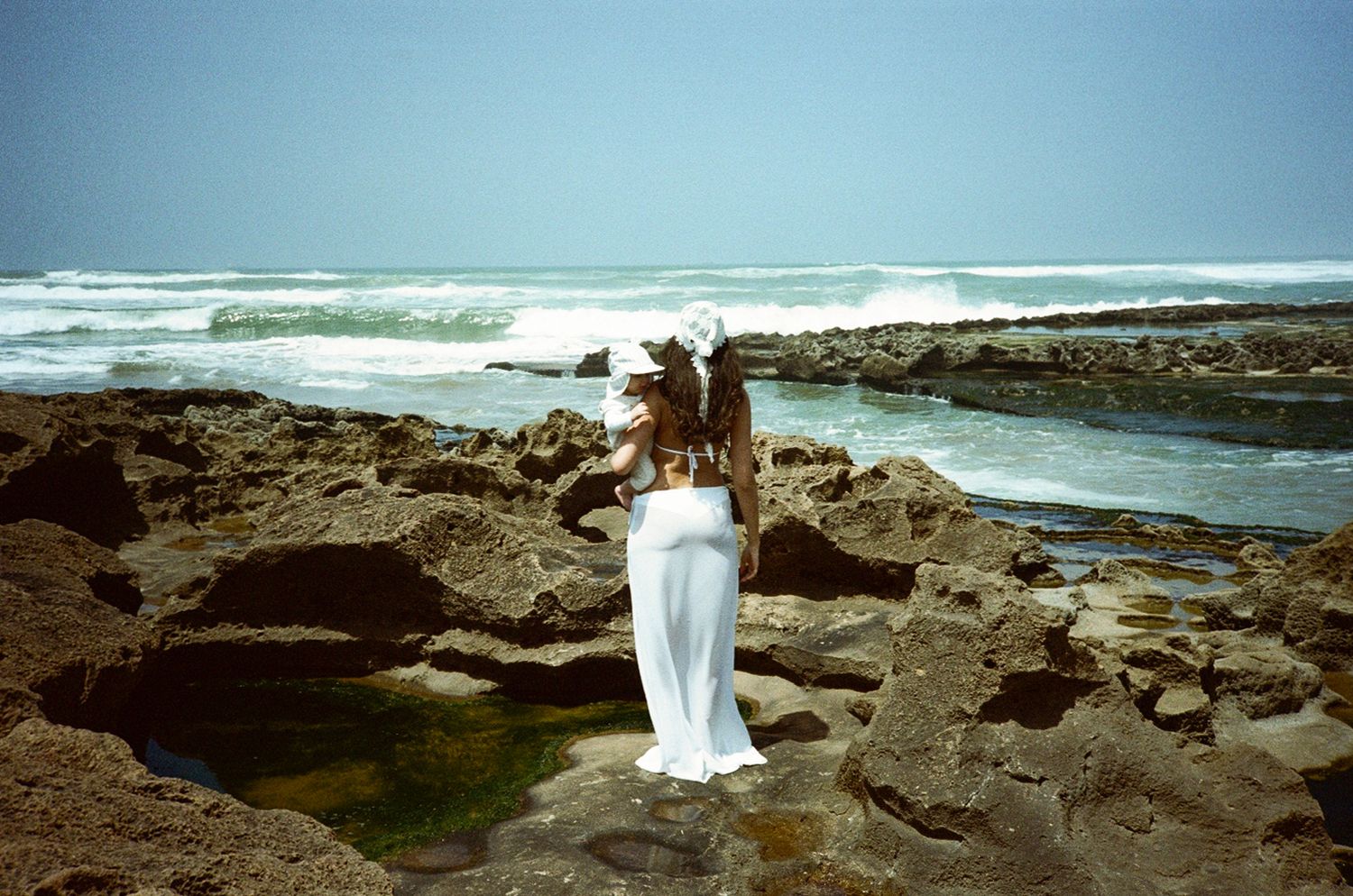 Person in white dress standing on rocky coastline with ocean waves and blue sky in background.