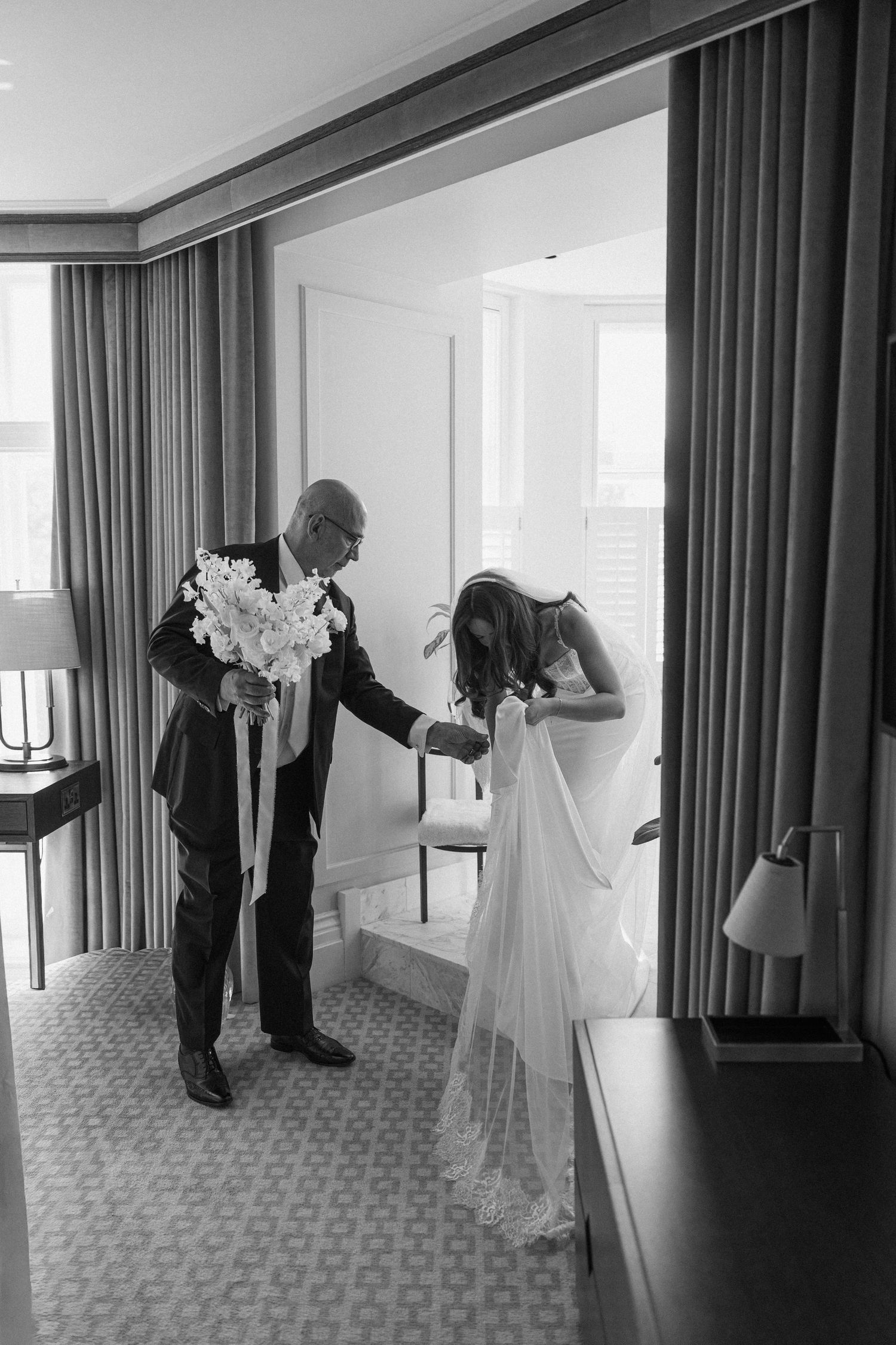 Black and white photo of a bride adjusting her veil while a man in a suit holds her bouquet of white flowers in a hotel room.