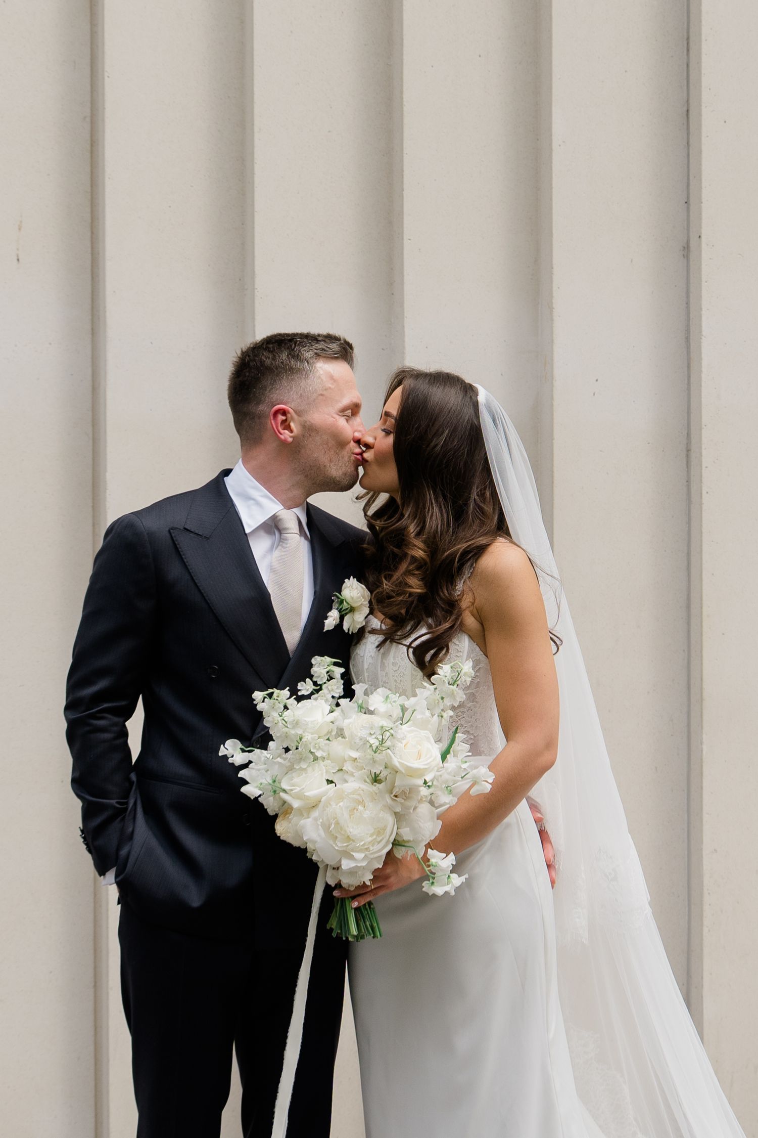 Bride and groom sharing a romantic kiss while bride holds white rose bouquet against neutral backdrop.