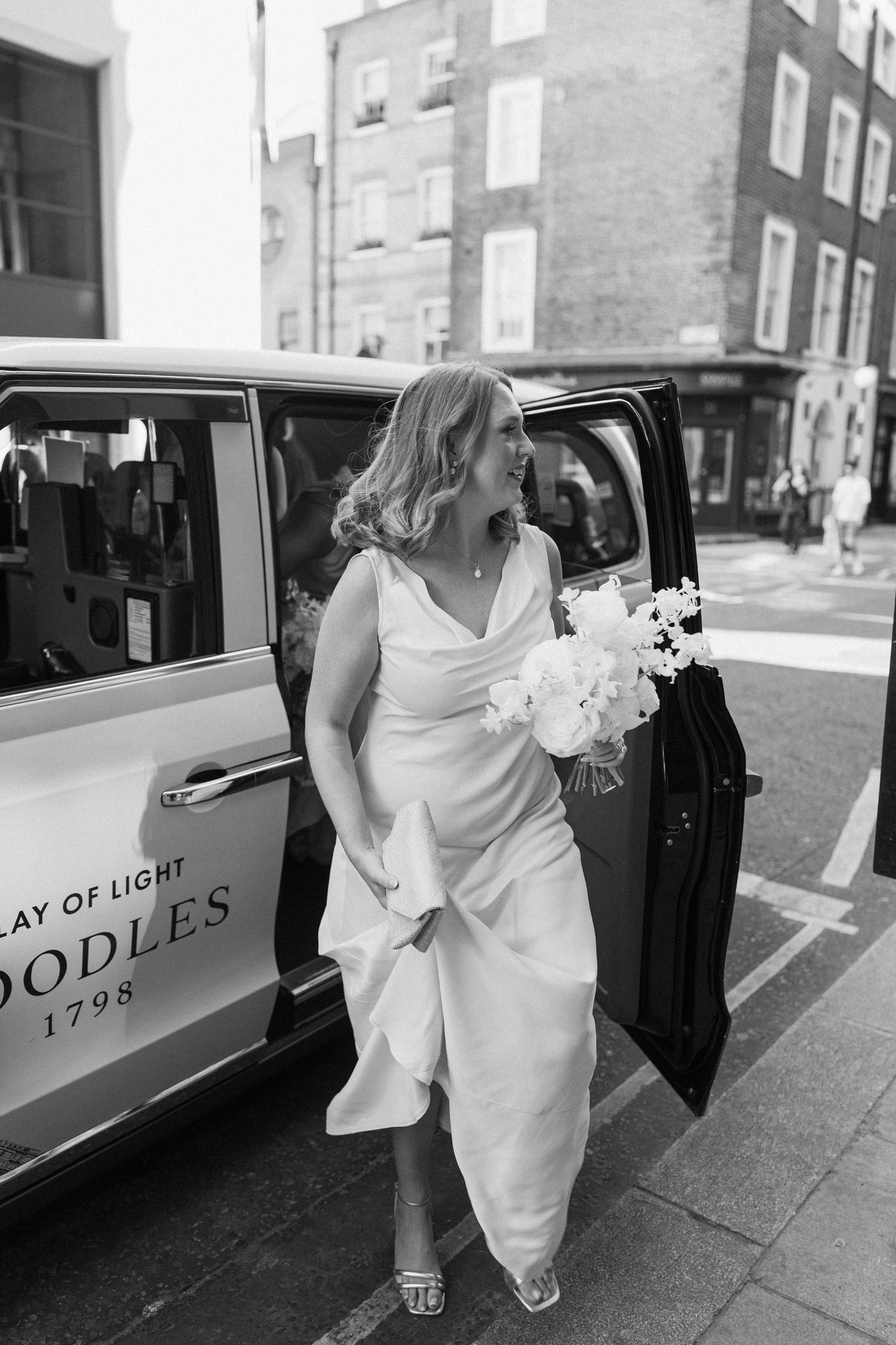 Bride in white gown holding bouquet stepping out of black London taxi cab on city street, black and white photo.