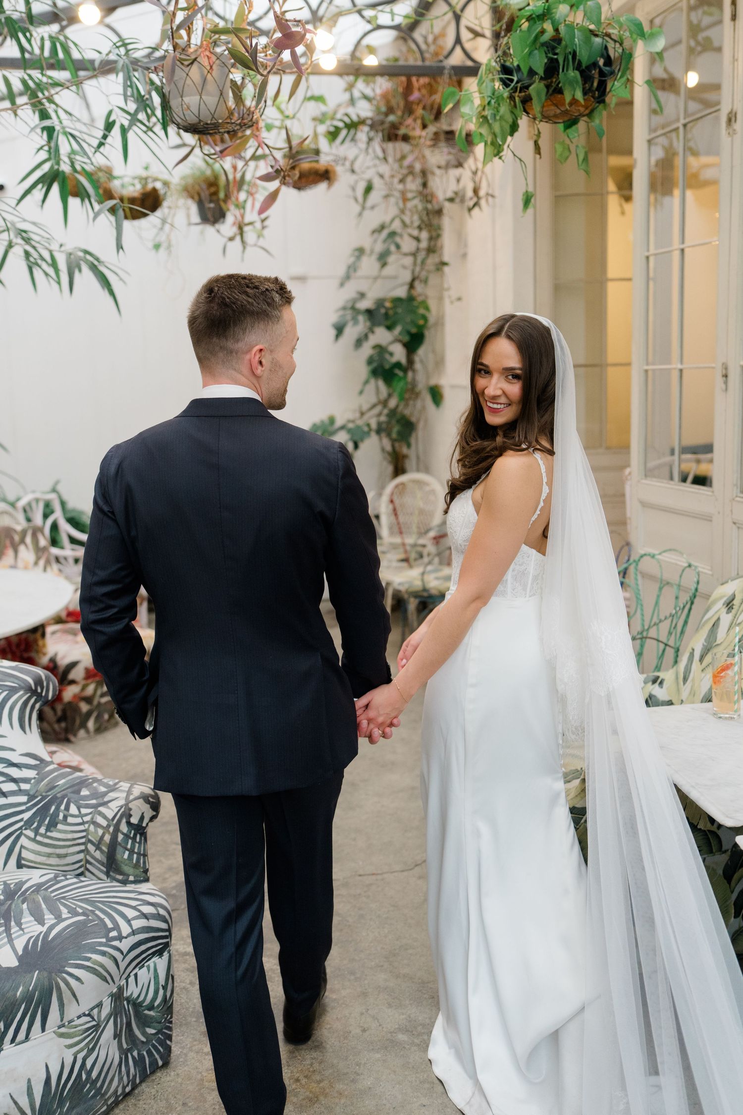 Bride in white gown with veil holds grooms hand walking through lush botanical wedding venue.