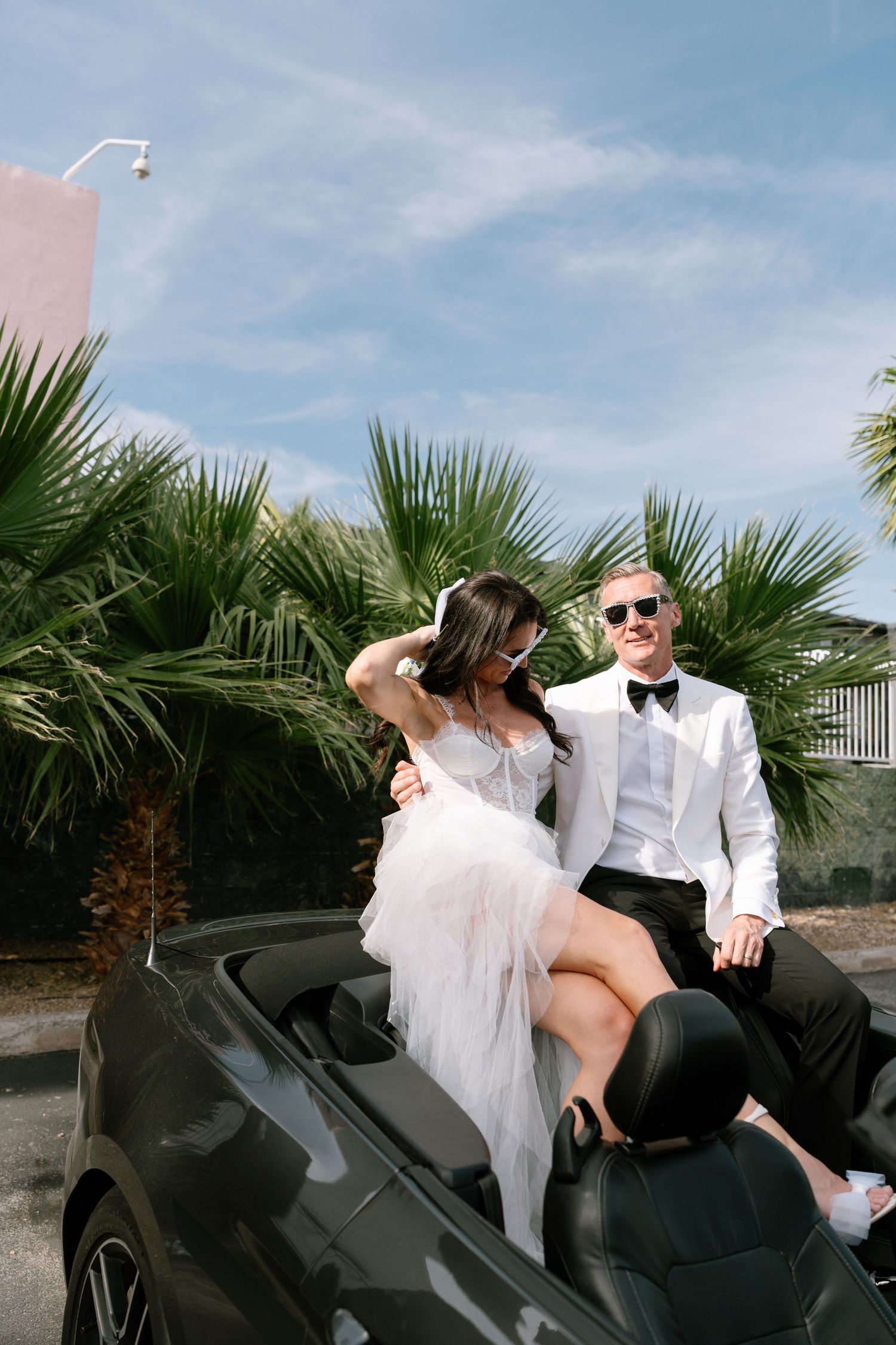 Newlyweds pose on black motorcycle in formal wedding attire with palm trees in background.