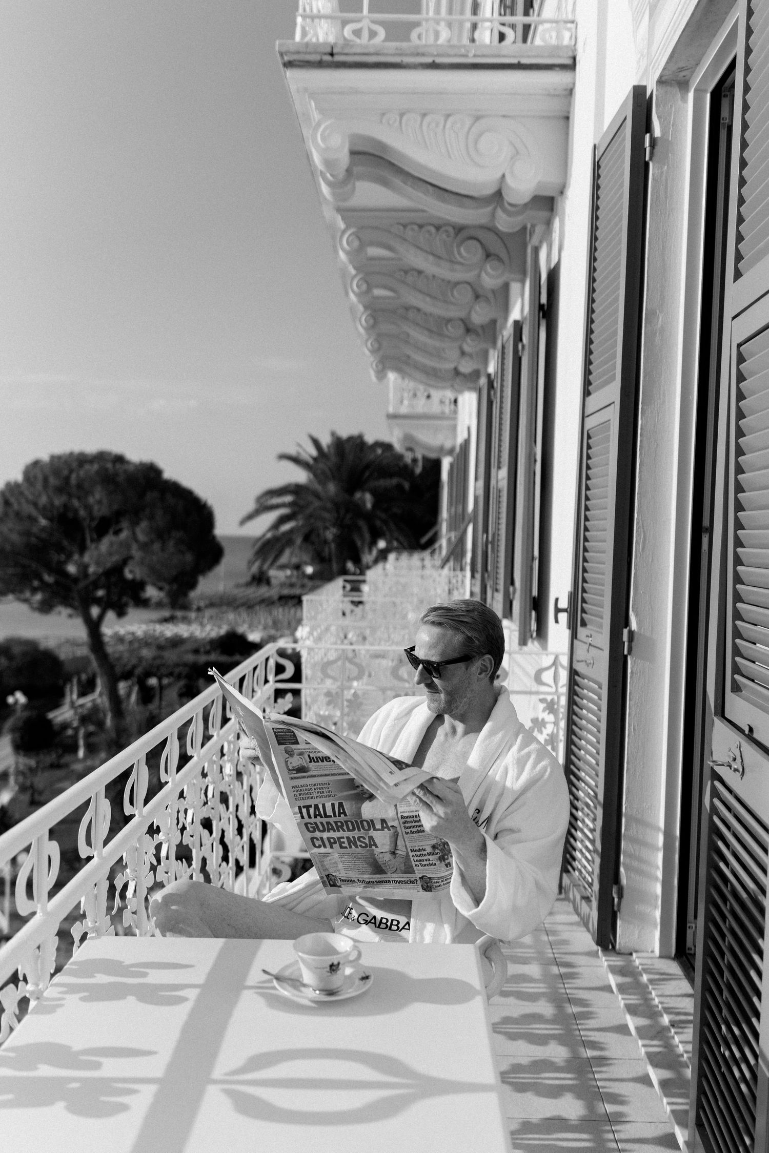 Black and white photo of a person reading an Italian newspaper on a sunny balcony with coffee, wearing sunglasses and a robe.
