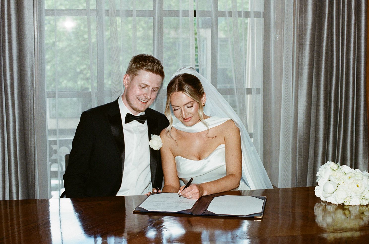 Bride and groom signing wedding register at wooden table, bride in strapless gown with veil, groom in black tuxedo.
