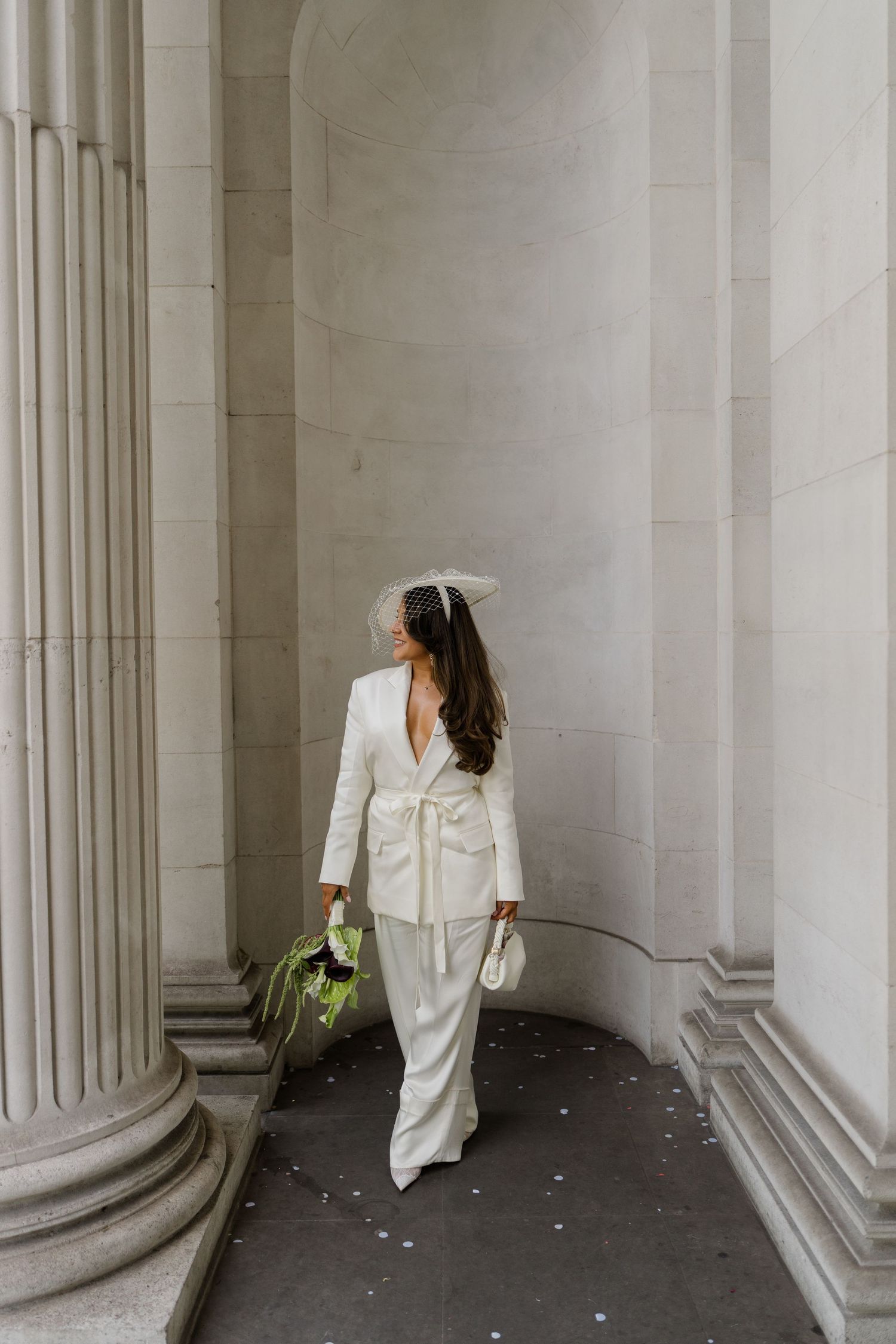 Bride in white backless suit and veil hat walks among stone columns at wedding venue.