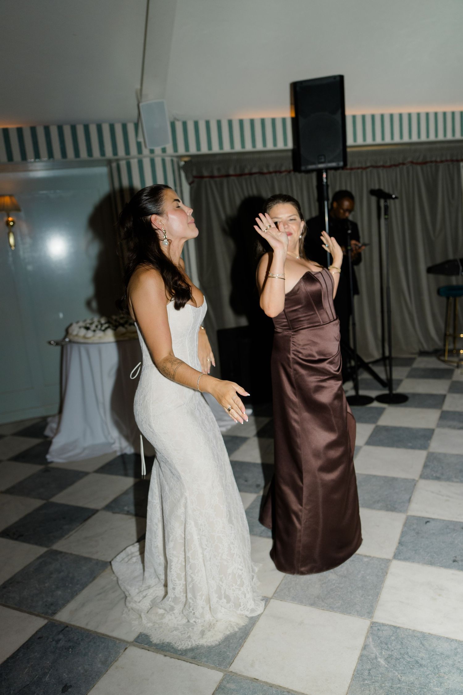 Bride in fitted lace gown dancing with bridesmaid in brown satin dress on checkered ballroom floor.