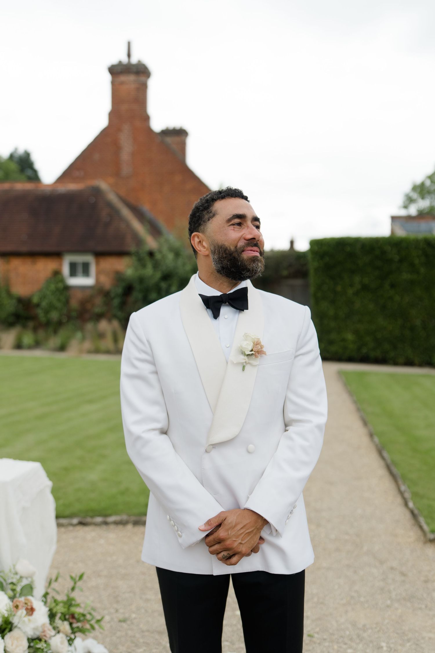 Groom in white tuxedo jacket and black bow tie smiling outdoors at English manor wedding venue.