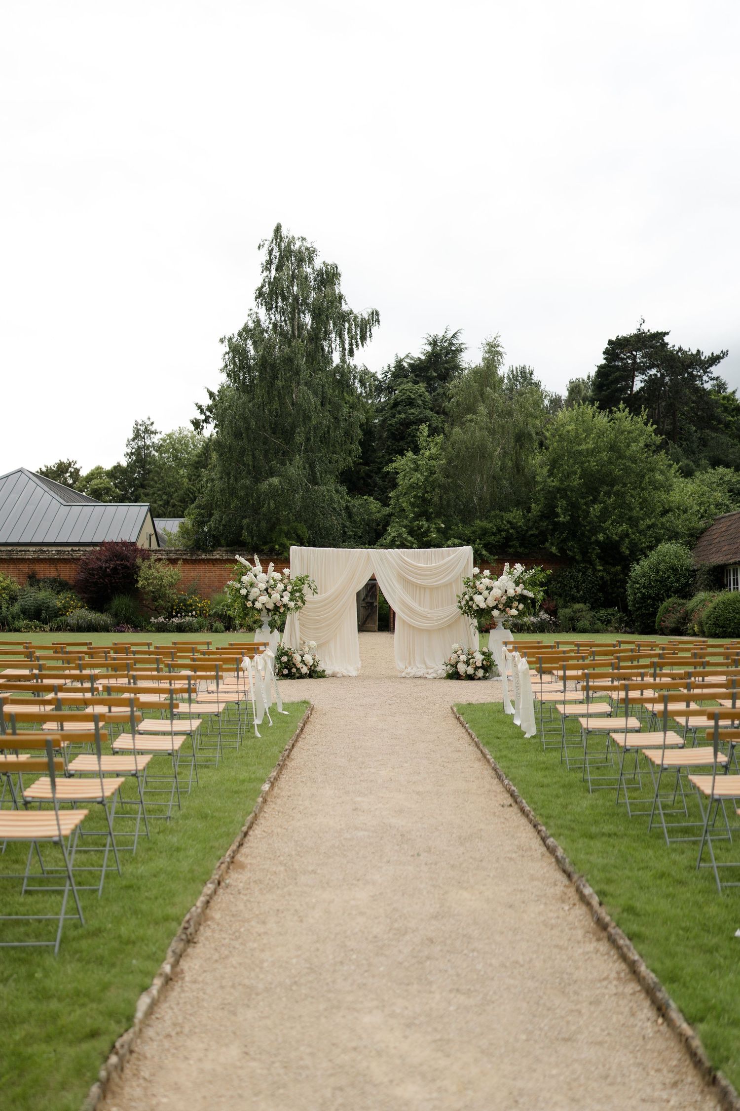 Outdoor wedding ceremony setup with white draped arch, floral arrangements, and rows of chairs on gravel aisle.