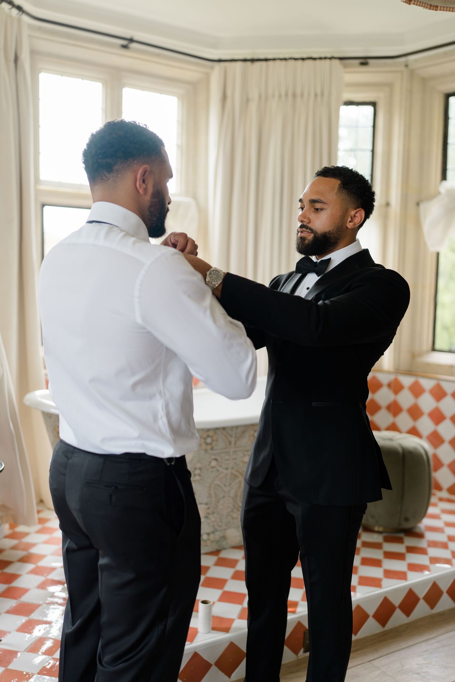Groomsman in tuxedo helping groom adjust collar in elegant red diamond-tiled bathroom before wedding.