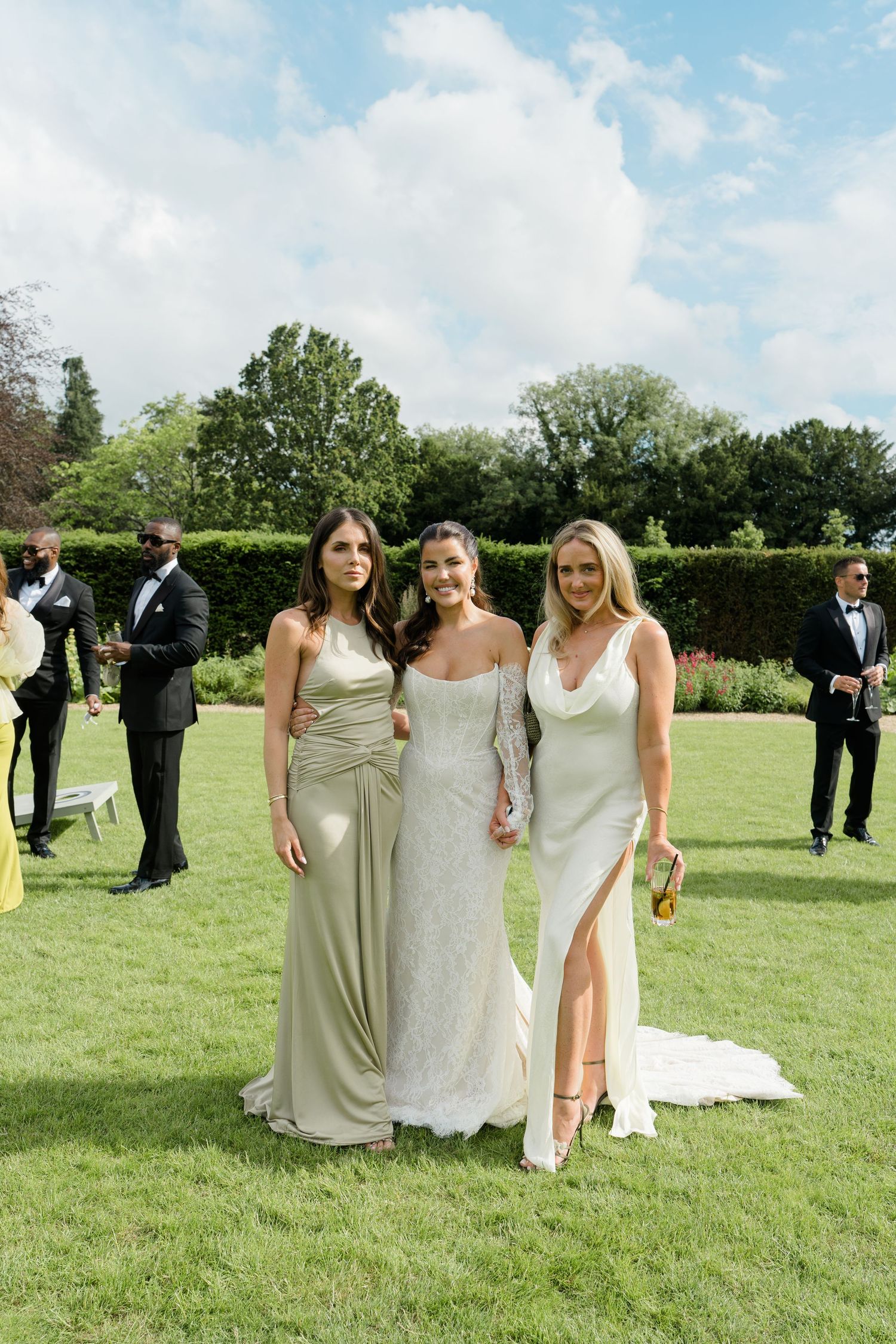 Bride in lace gown poses with two bridesmaids in sage green satin dresses on sunny garden lawn at elegant wedding.