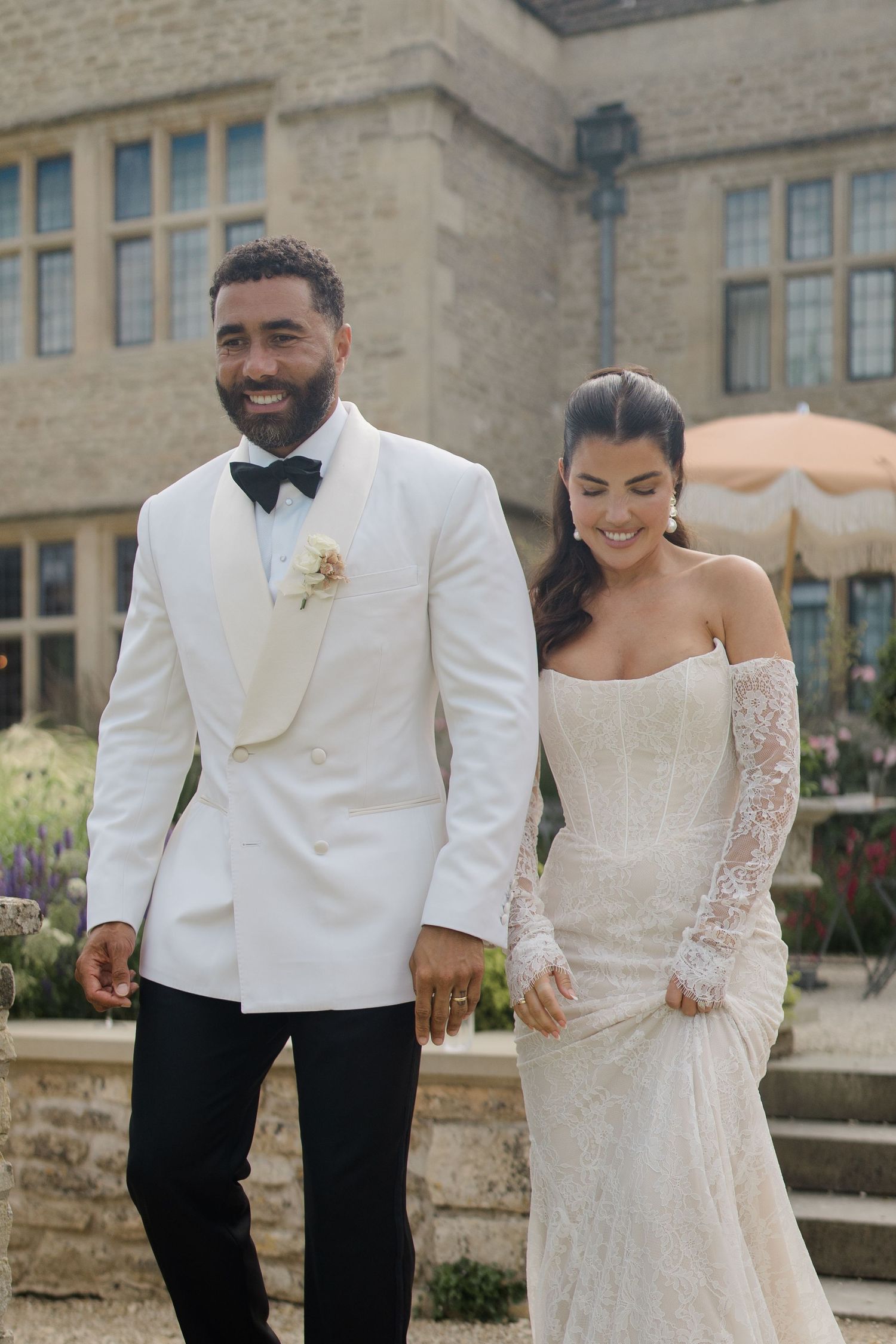 Groom in white tuxedo jacket walking with bride in lace off-shoulder gown at manor.