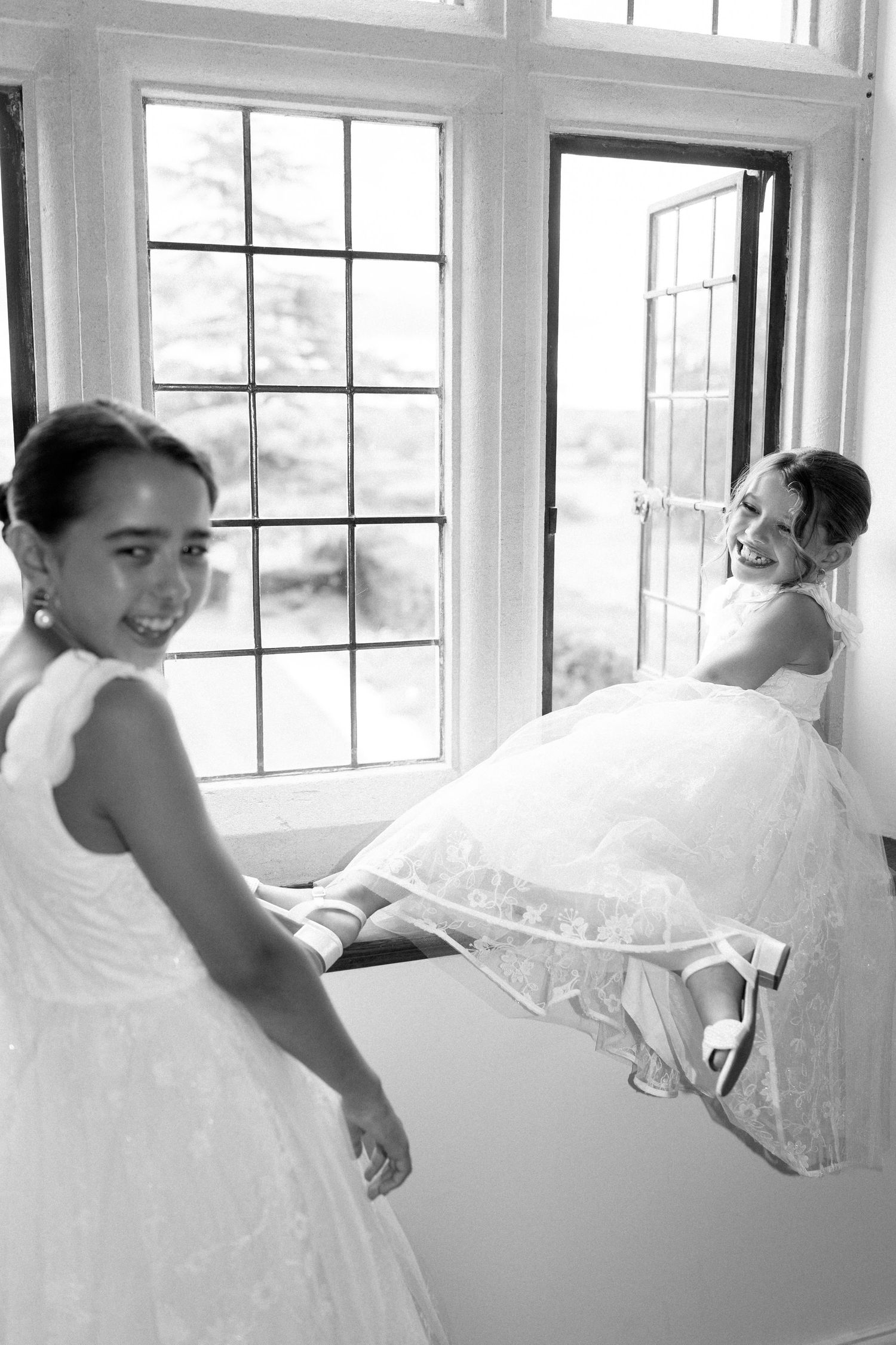 Black and white photo of two flower girls in white dresses laughing by a window.