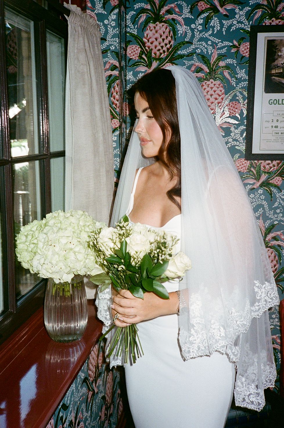 Bride in white strapless gown and lace veil holds white hydrangea bouquet by window with floral wallpaper.