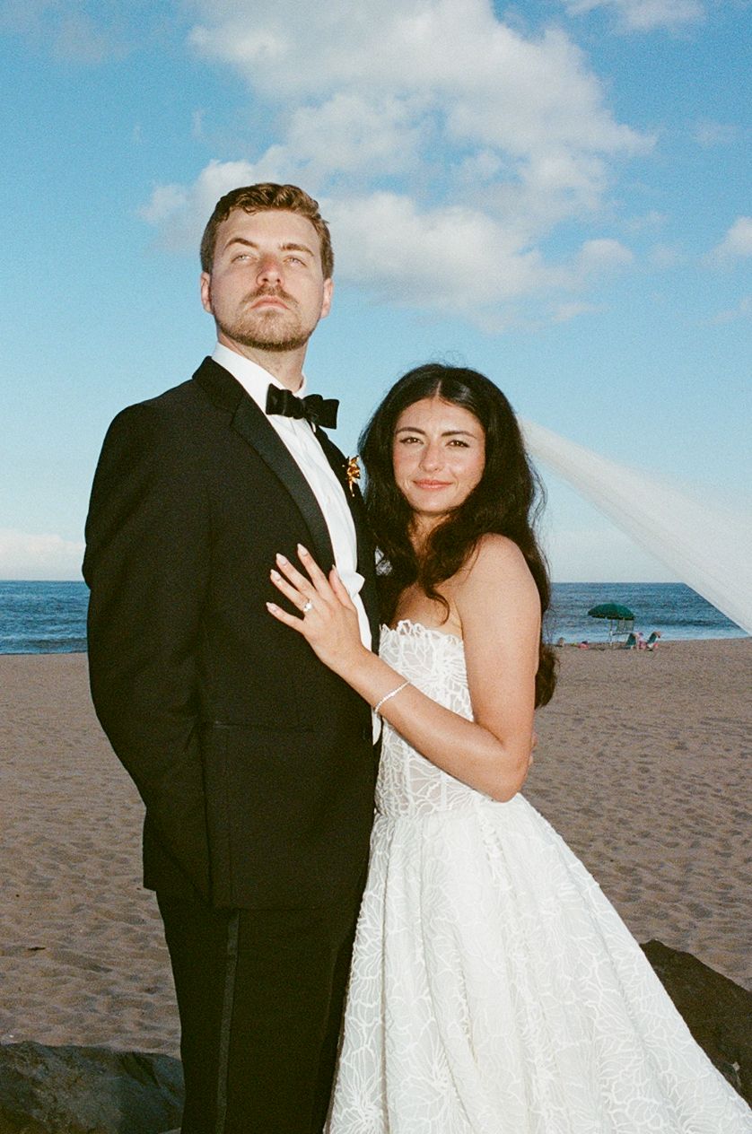 Bride and groom pose on a sandy beach, she in a white gown and he in a black tuxedo with bow tie.