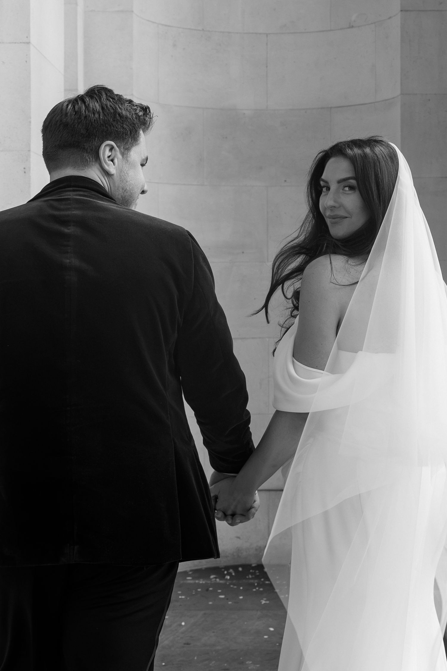 Black and white photo of a bride in an off-the-shoulder gown and veil glancing back while holding hands with a groom in a velvet jacket.
