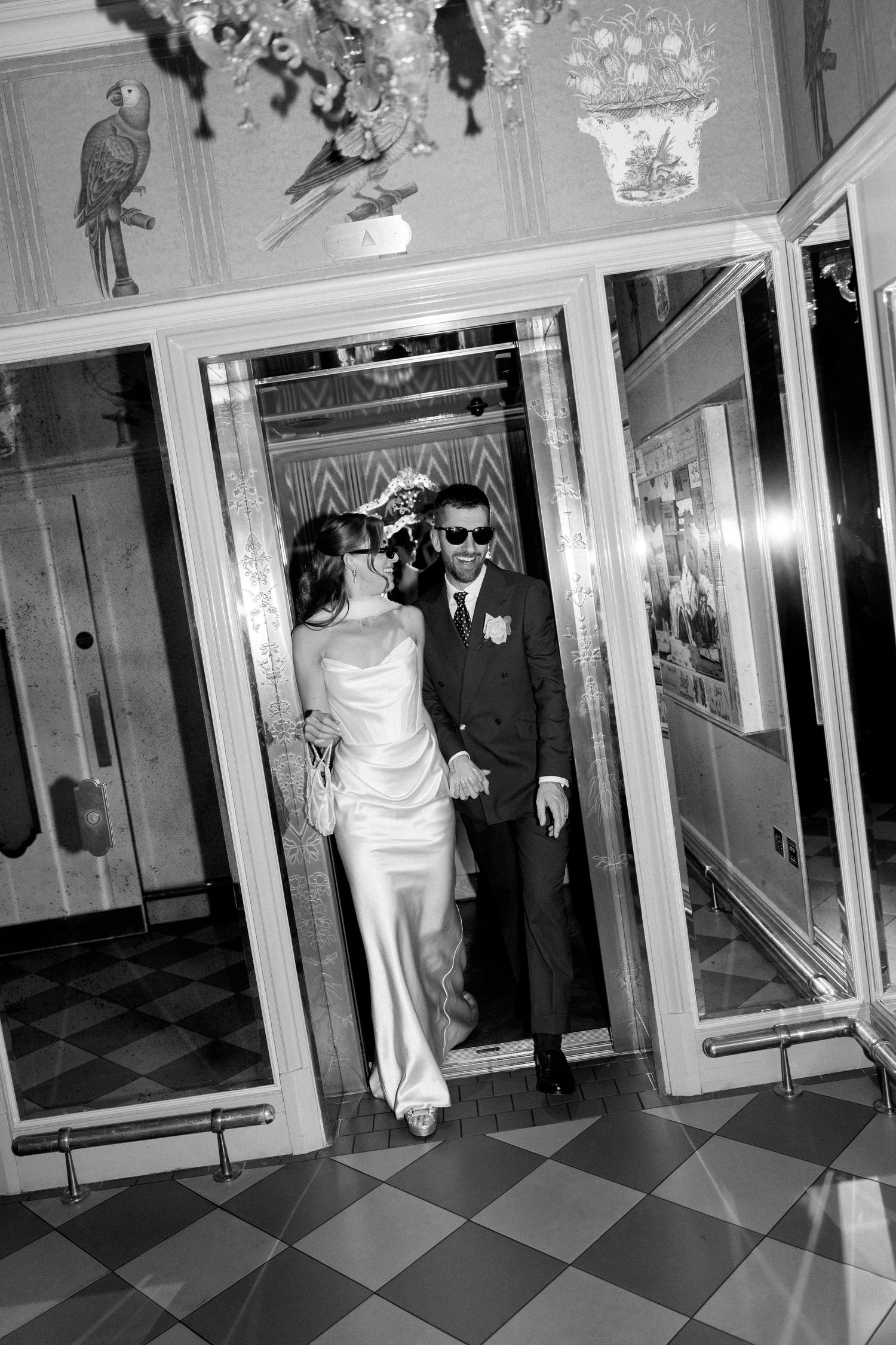 Black and white photo of bride and groom in sunglasses exiting ornate mirrored elevator together.