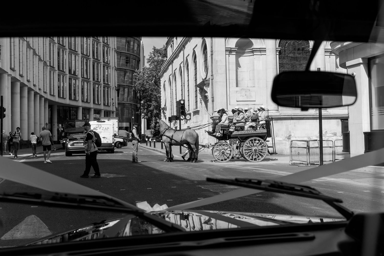 Black and white view through car windshield of horse-drawn carriage on a city street.