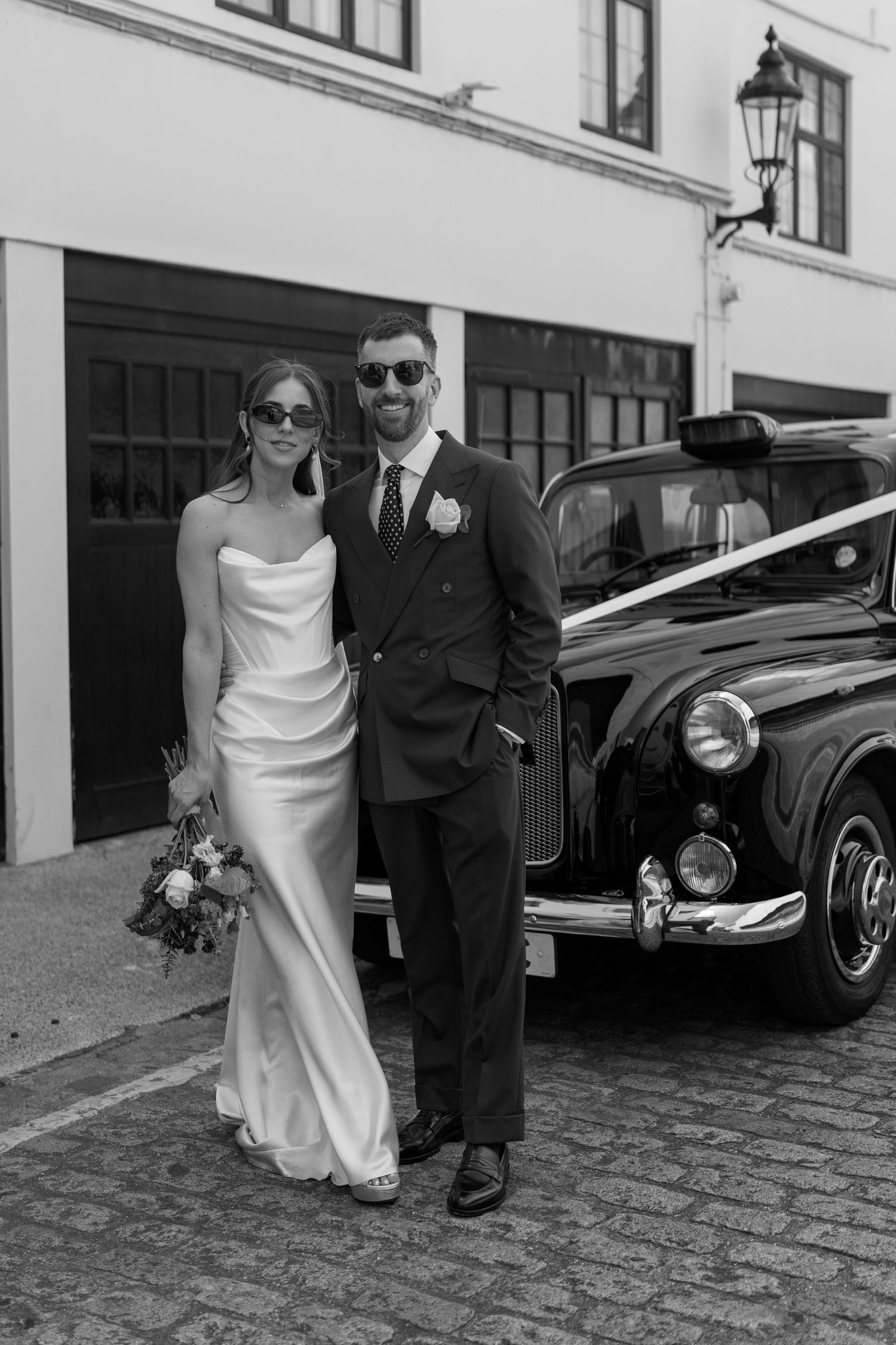 Bride and groom in sunglasses pose beside vintage black London taxi cab on cobblestone street.