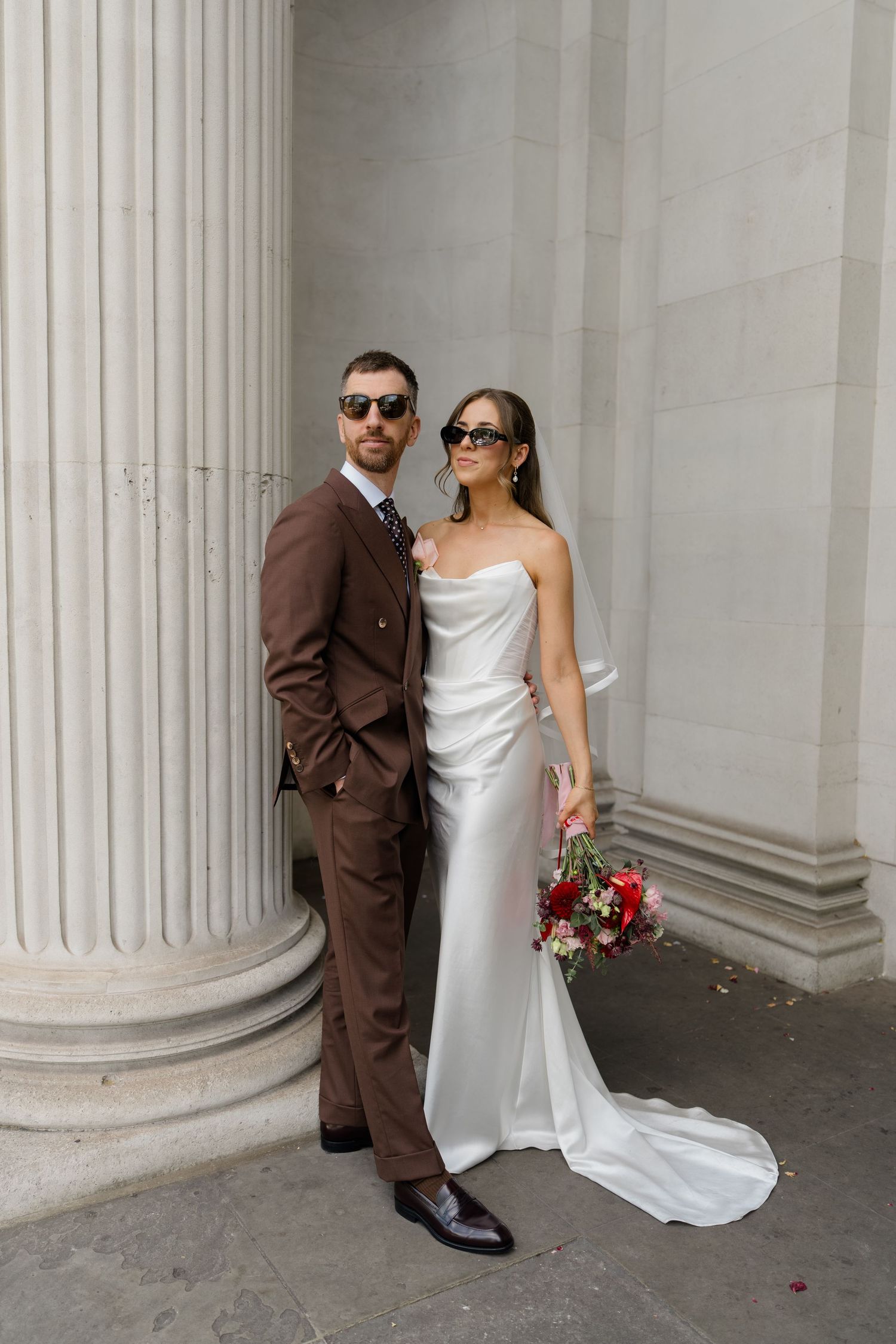Stylish bride and groom in sunglasses pose by stone columns, she wears a white satin gown holding a red bouquet.