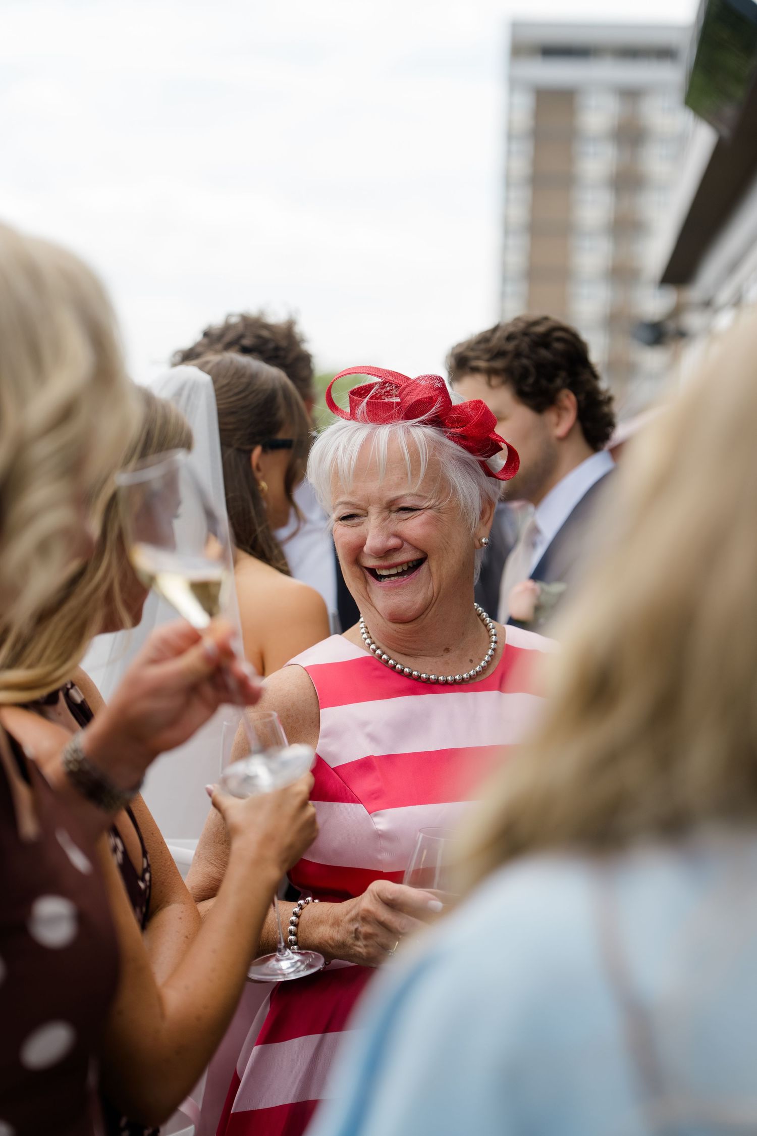Joyful wedding guest in a red fascinator laughs while holding champagne at an outdoor reception.